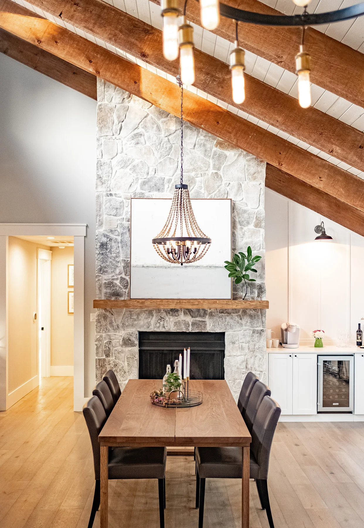Bright dining room with a stone fireplace, wood beams, and a hanging chandelier.