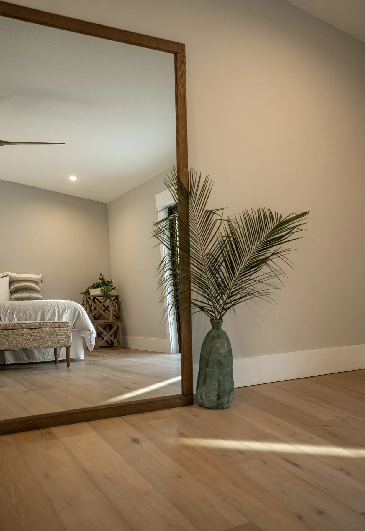Quiet bedroom interior with a woven bedside table and a tall glass vase holding palm fronds.