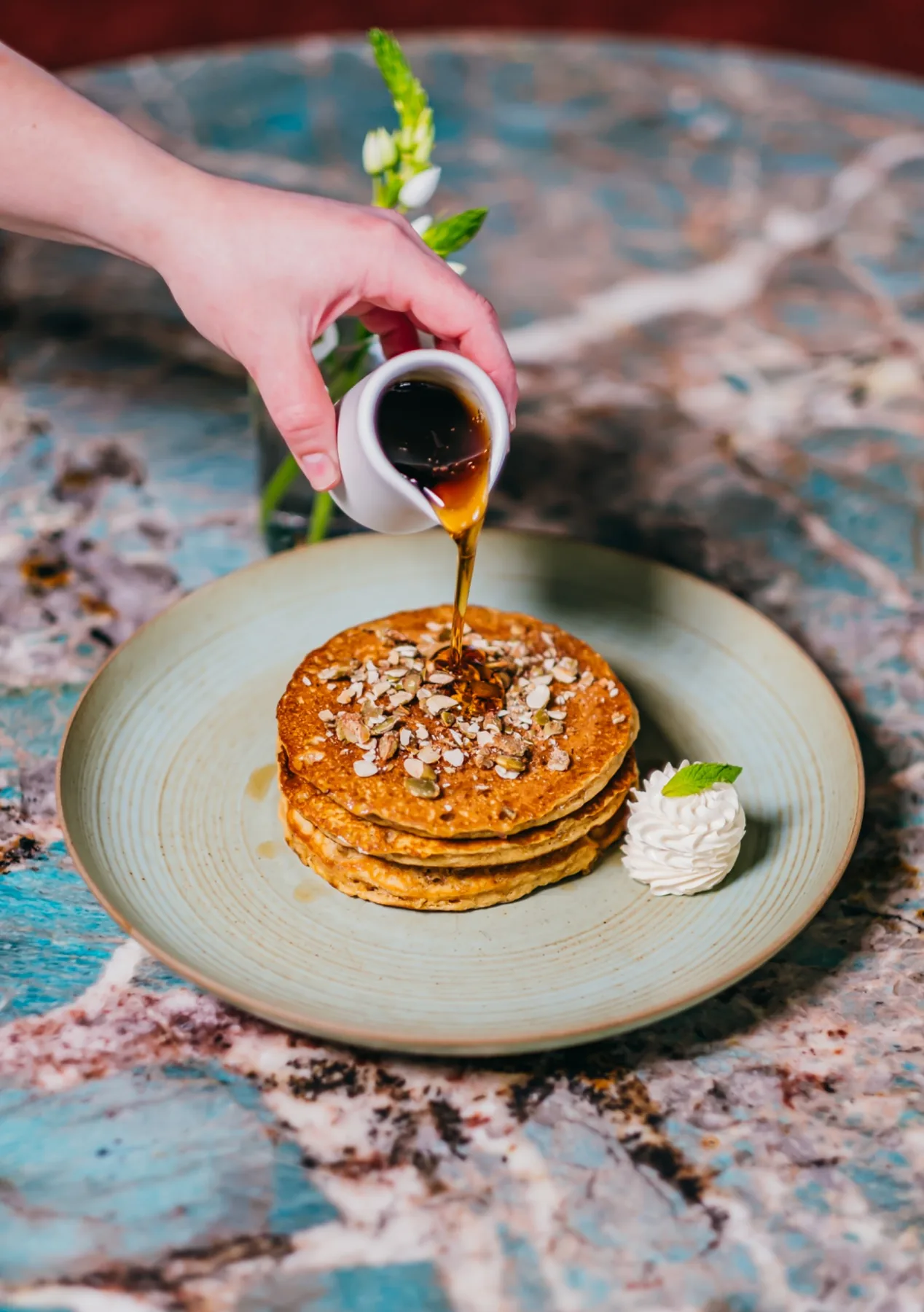 Stack of pancakes with pecans as syrup pours from a small pitcher.