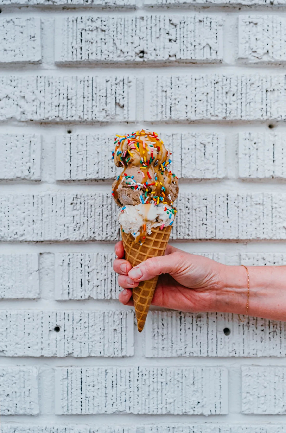 Hand holding a sprinkle-topped ice cream cone against a white brick wall.