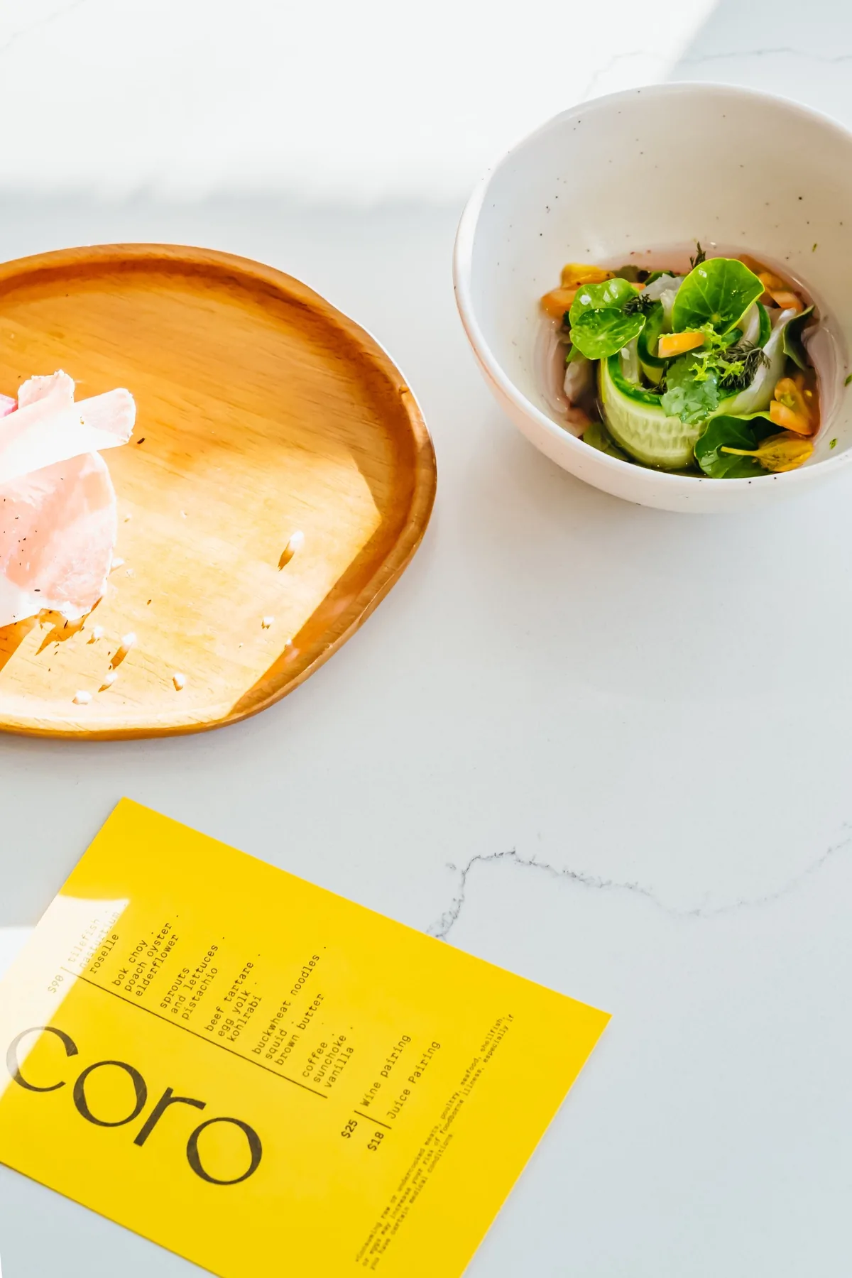 Overhead table setting with a plated dish and a bright yellow Coro menu.