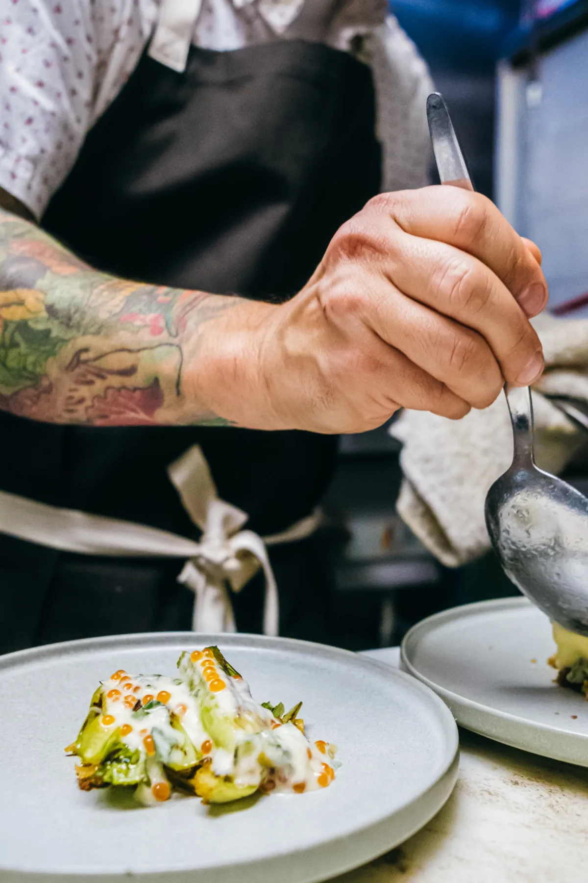 Tattooed hand ladles sauce over a dish in the kitchen.