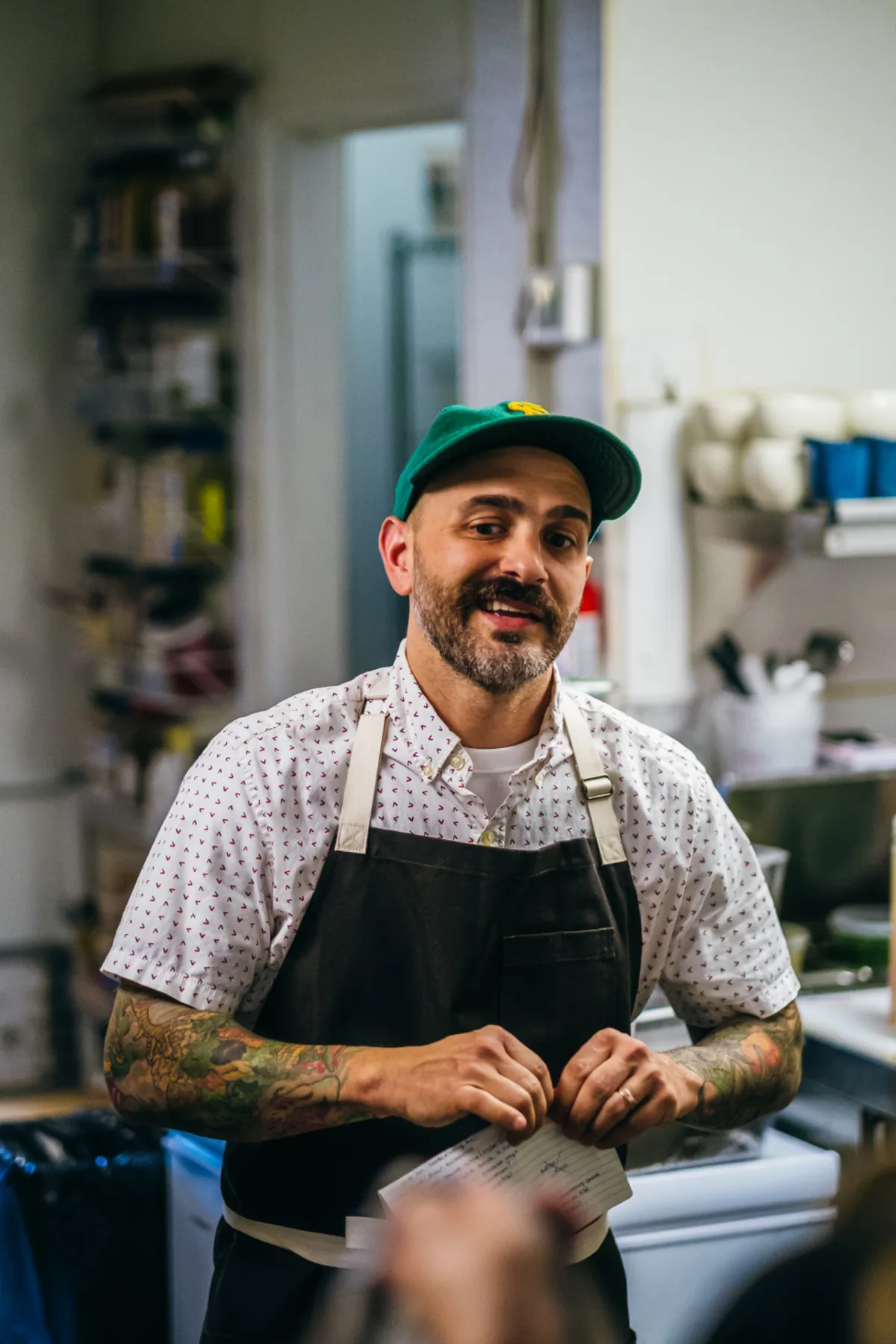 Chef in a green cap and dark apron looking across the kitchen.