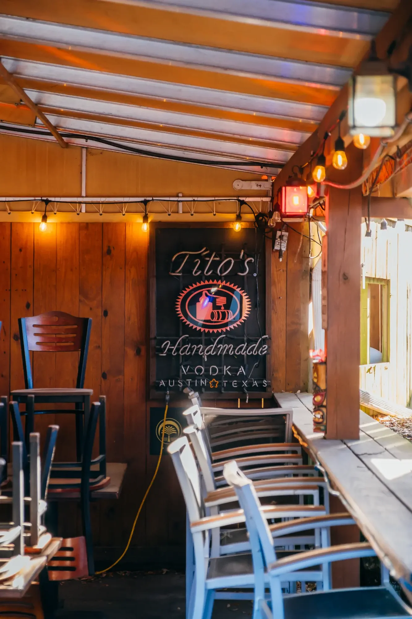 Covered wooden patio with stacked chairs warm string lights and a neon sign.