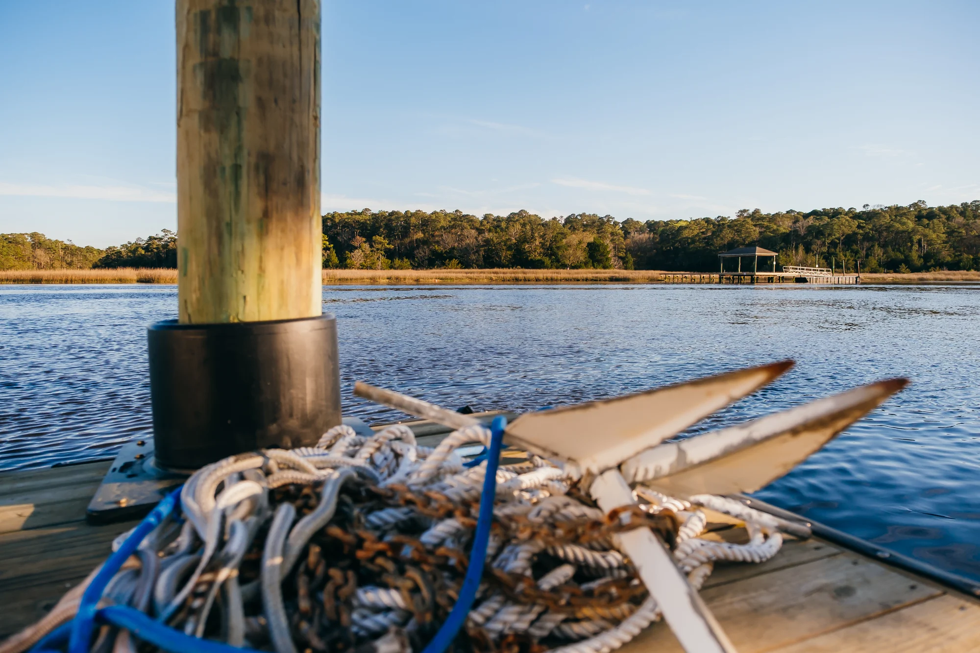 Weathered dock piling and coiled ropes in foreground with calm river and distant pier.