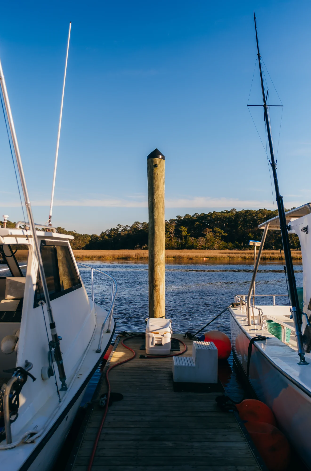 Sunlit wooden dock between two boats with central piling and calm blue water.
