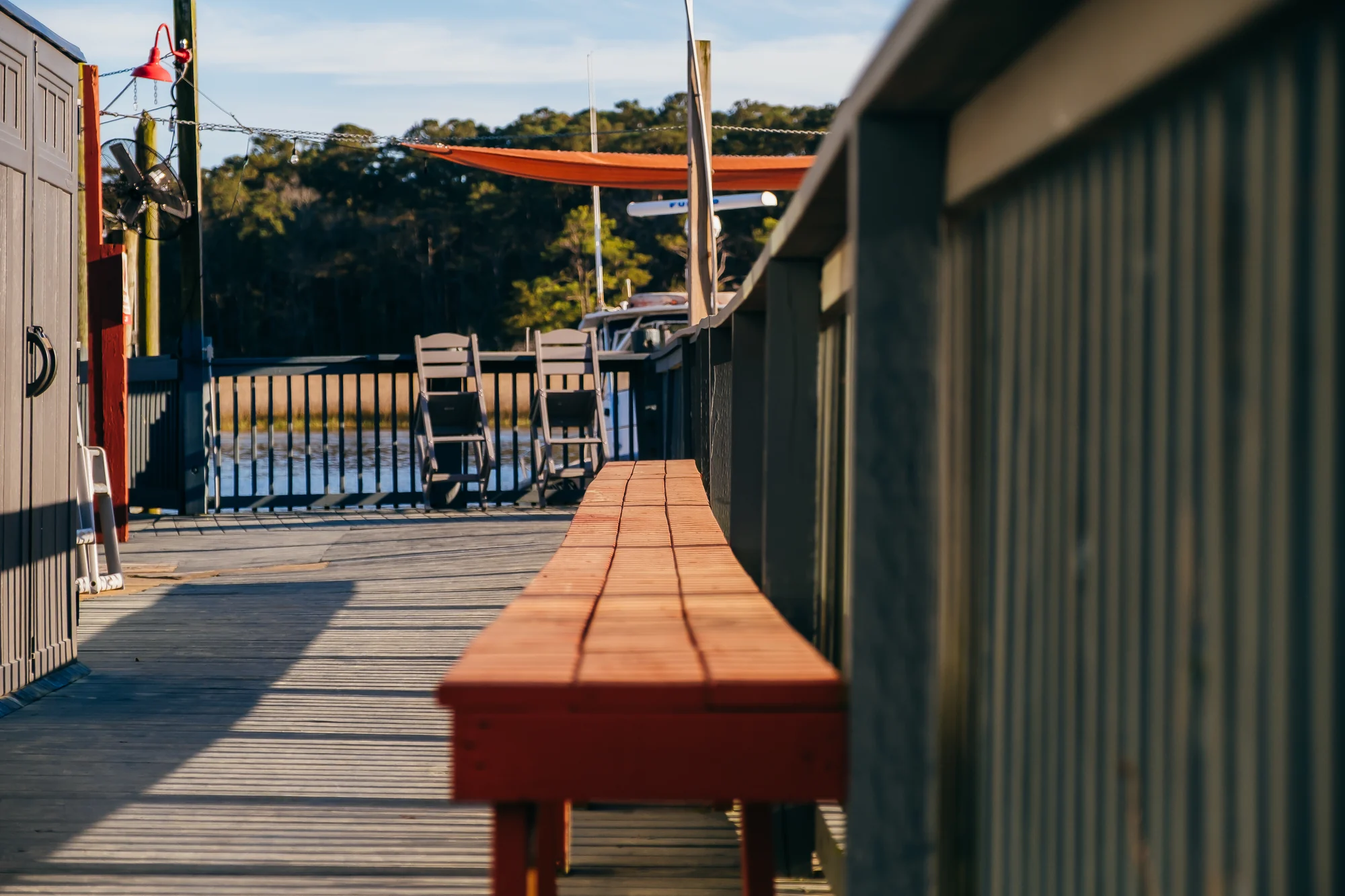 Wooden dock bench leading to two chairs and boat under orange canopy.
