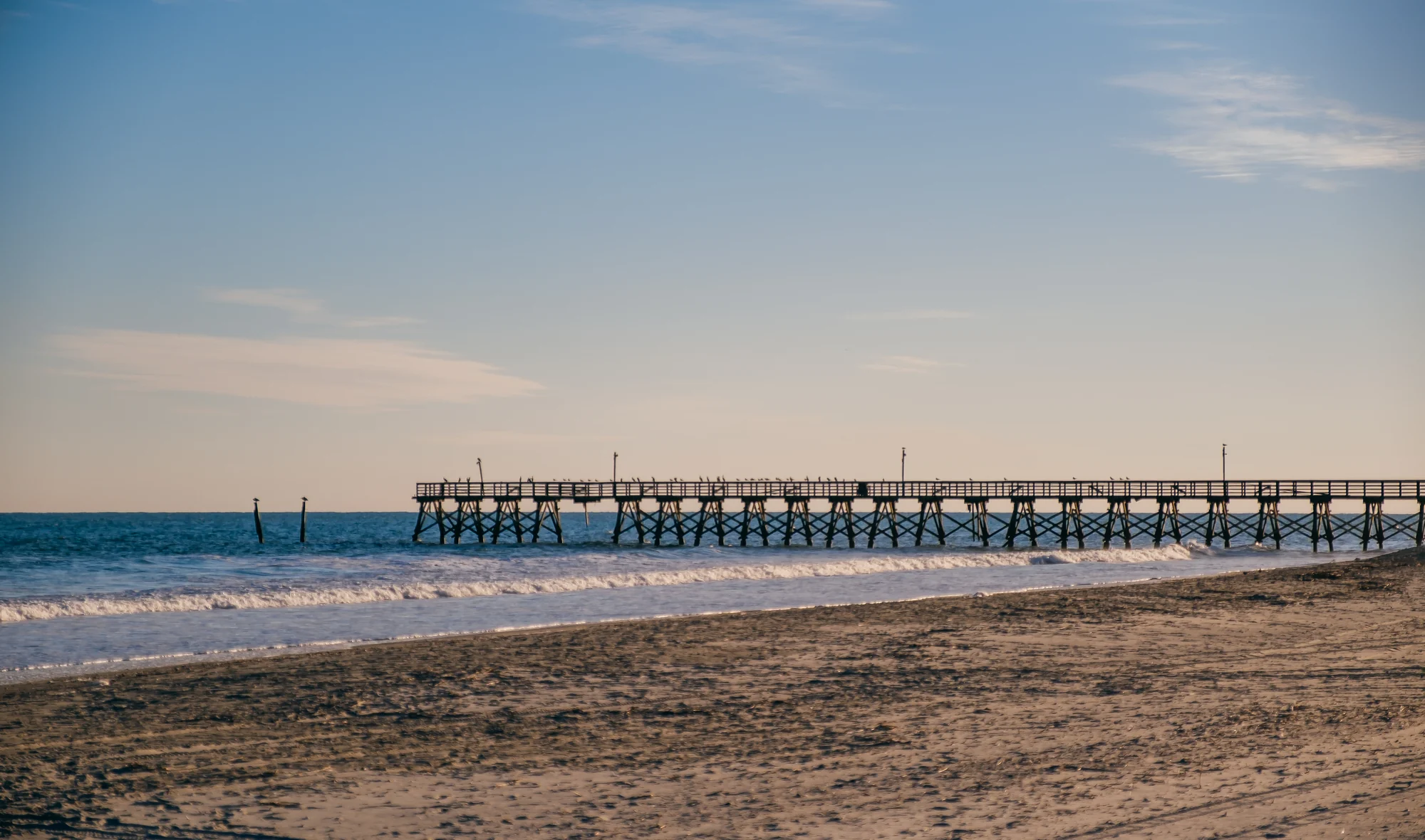 Wooden pier extending right across calm blue ocean with sandy foreground and pastel sky.