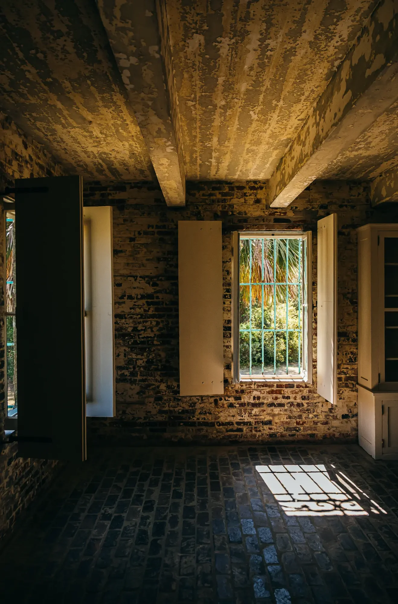 Peeling ceiling beams and brick walls frame barred window casting sunlight grid onto cobblestone floor.