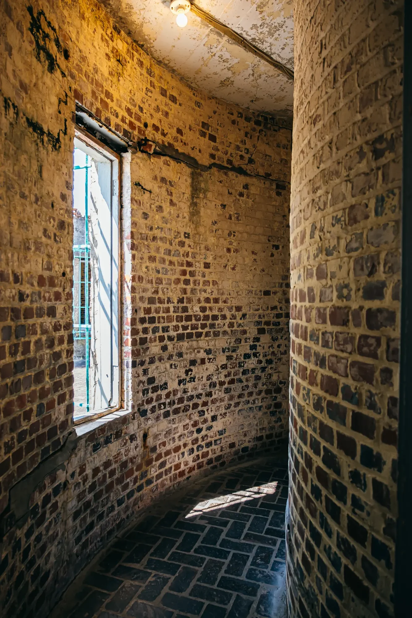 Curved aged brick stairwell lit by window and single exposed bulb casting shadow.