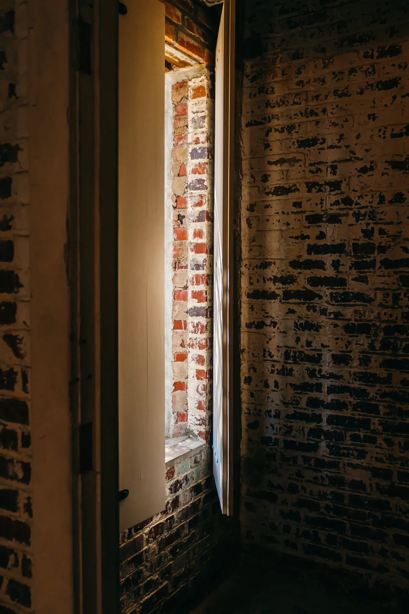 Narrow vertical shaft of warm light revealing exposed red brick and peeling painted wall.