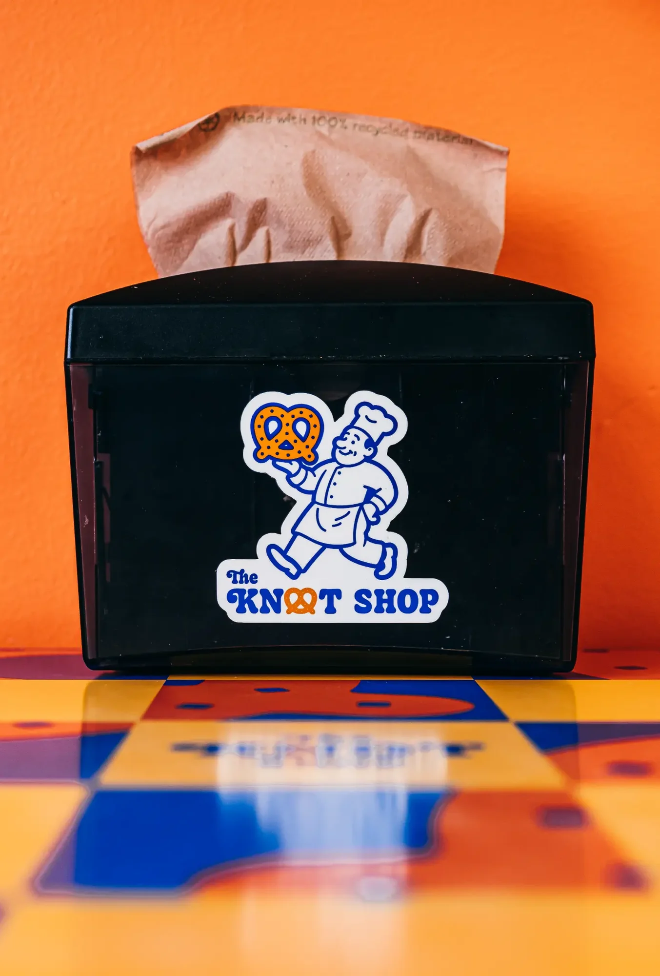 Black napkin dispenser with brown paper napkin against bright orange wall and colorful tabletop reflection.