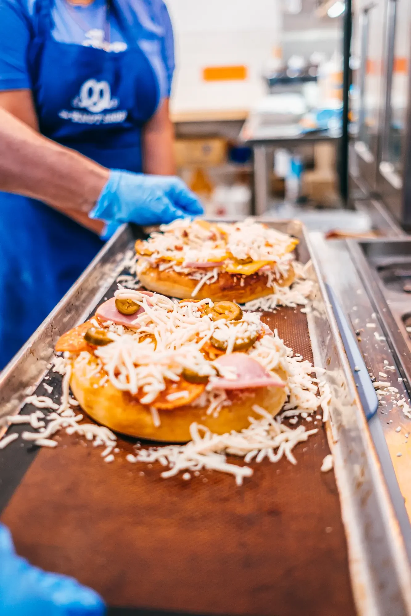 Tray of topped mini pizzas with shredded cheese and jalapeños held by gloved hand.