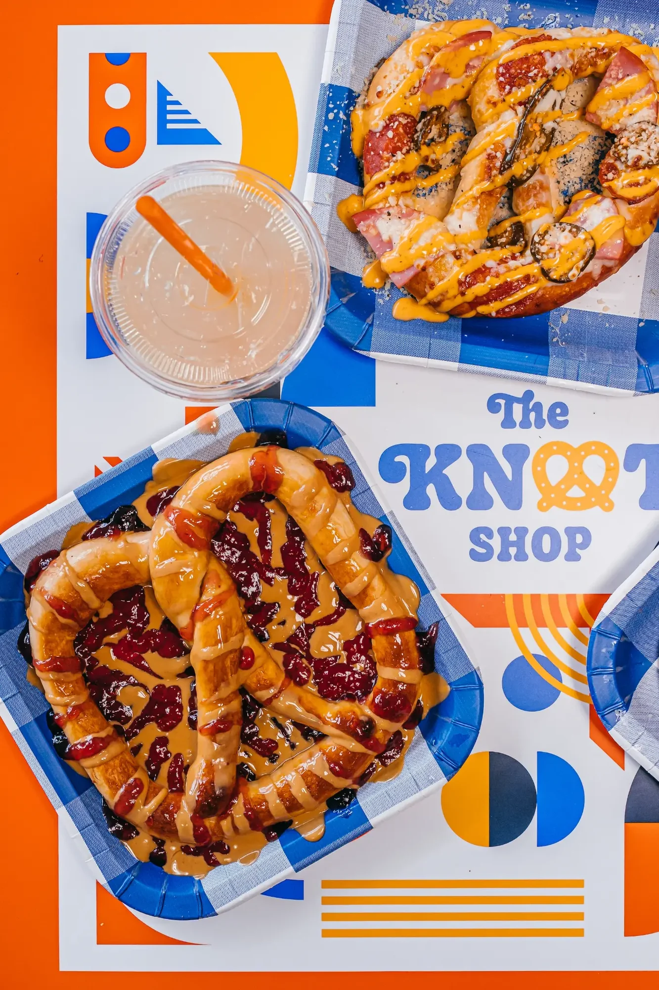 Sauced pretzels on blue checkered trays beside a plastic drink with orange straw.