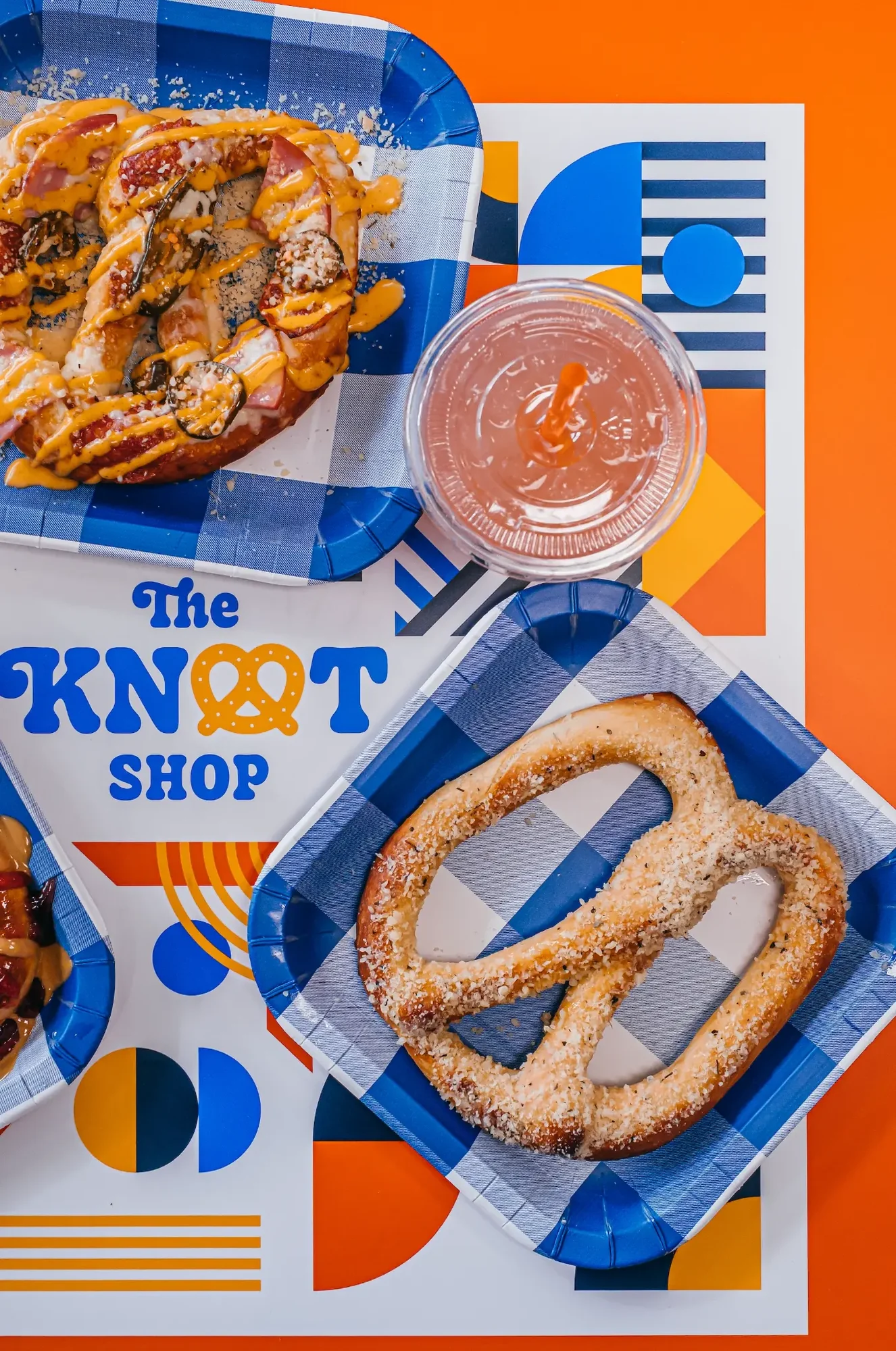 Salted pretzel and sauce drizzled pretzel beside pink drink on geometric orange background.