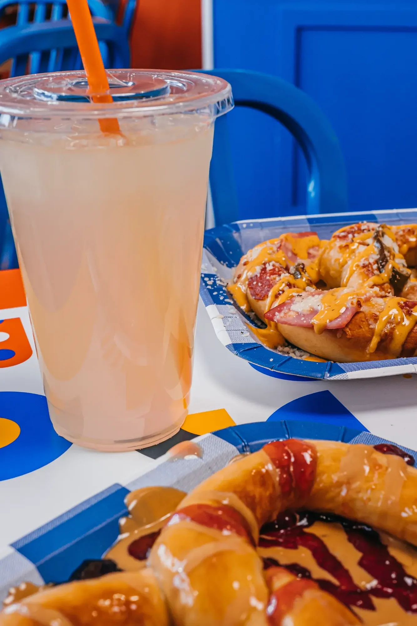 Tall plastic cup with pale pink drink and orange straw beside glazed donuts with sauces.