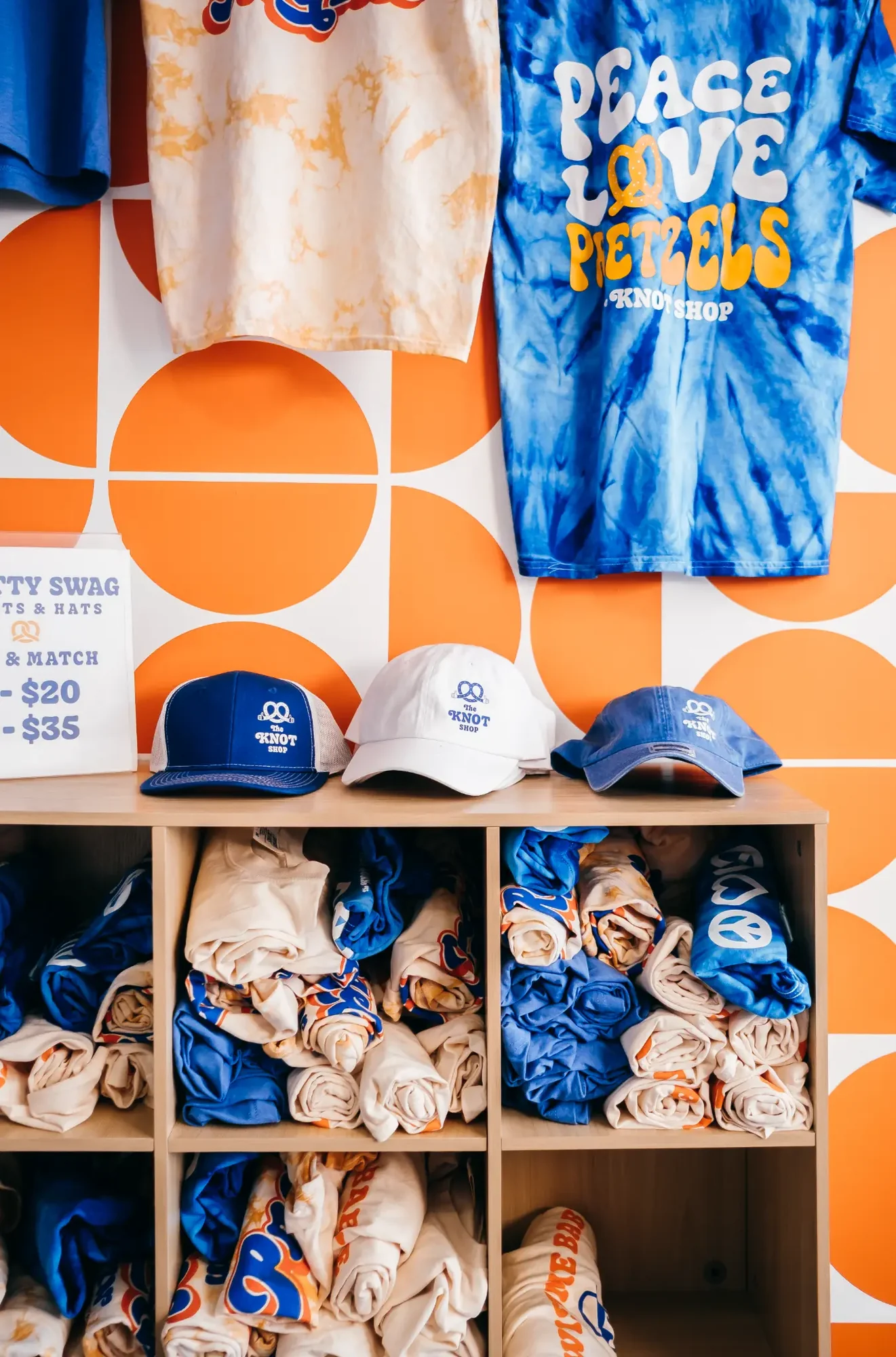 Blue tie dye shirt and caps displayed against orange geometric wall above cubbies of rolled shirts.