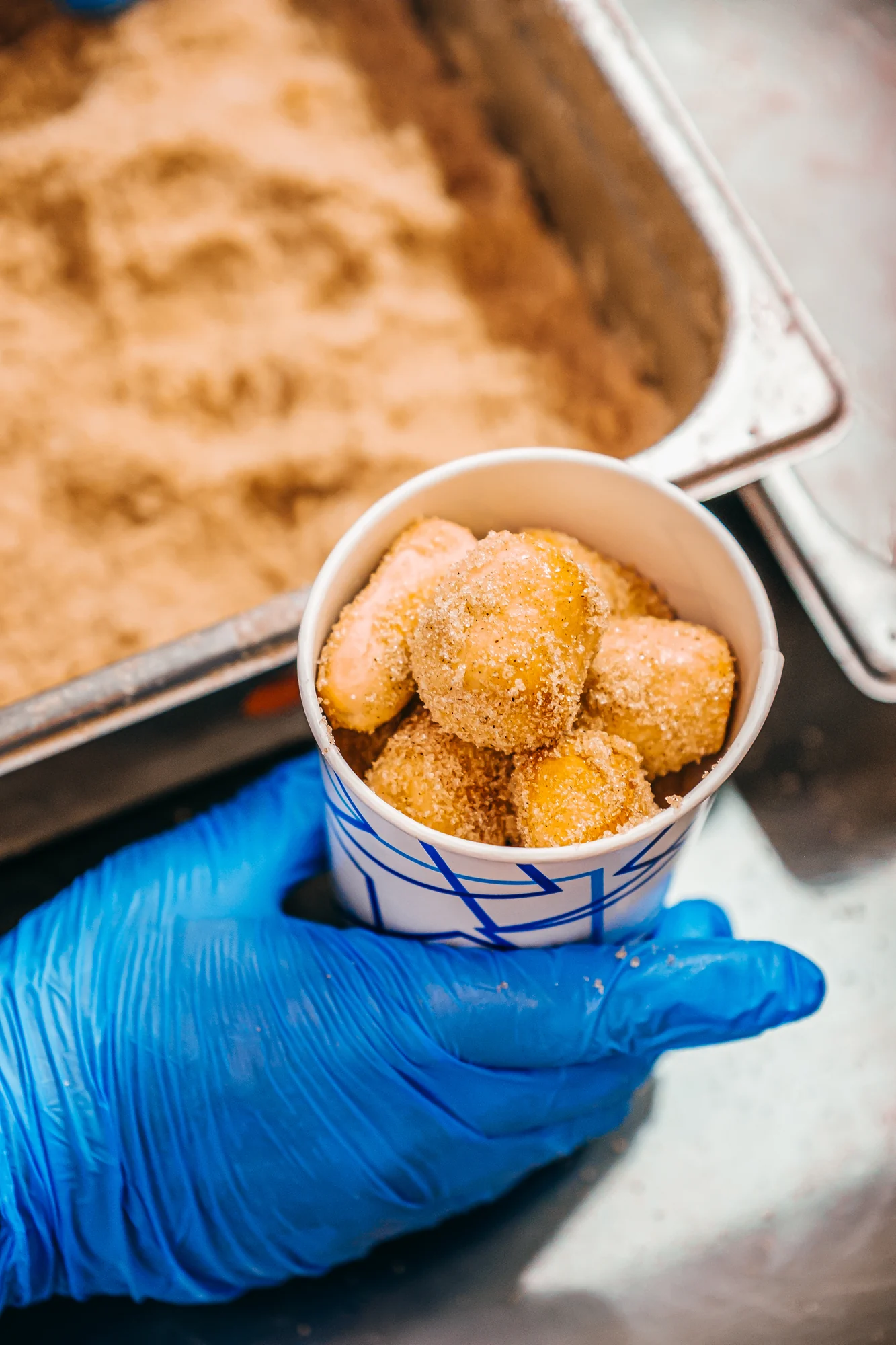 Blue gloved hand holding paper cup of sugar coated doughnut holes by metal tray.