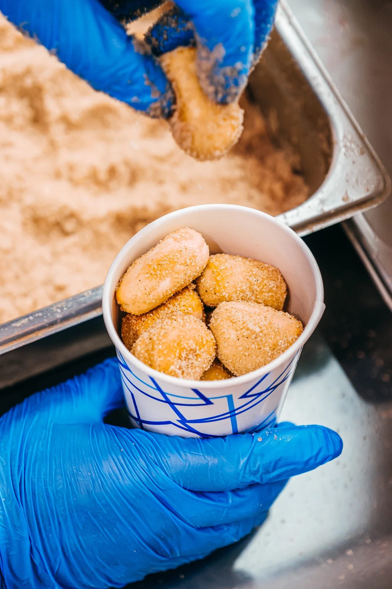 Blue gloved hand holding paper cup filled with sugar coated mini donuts under warm light.