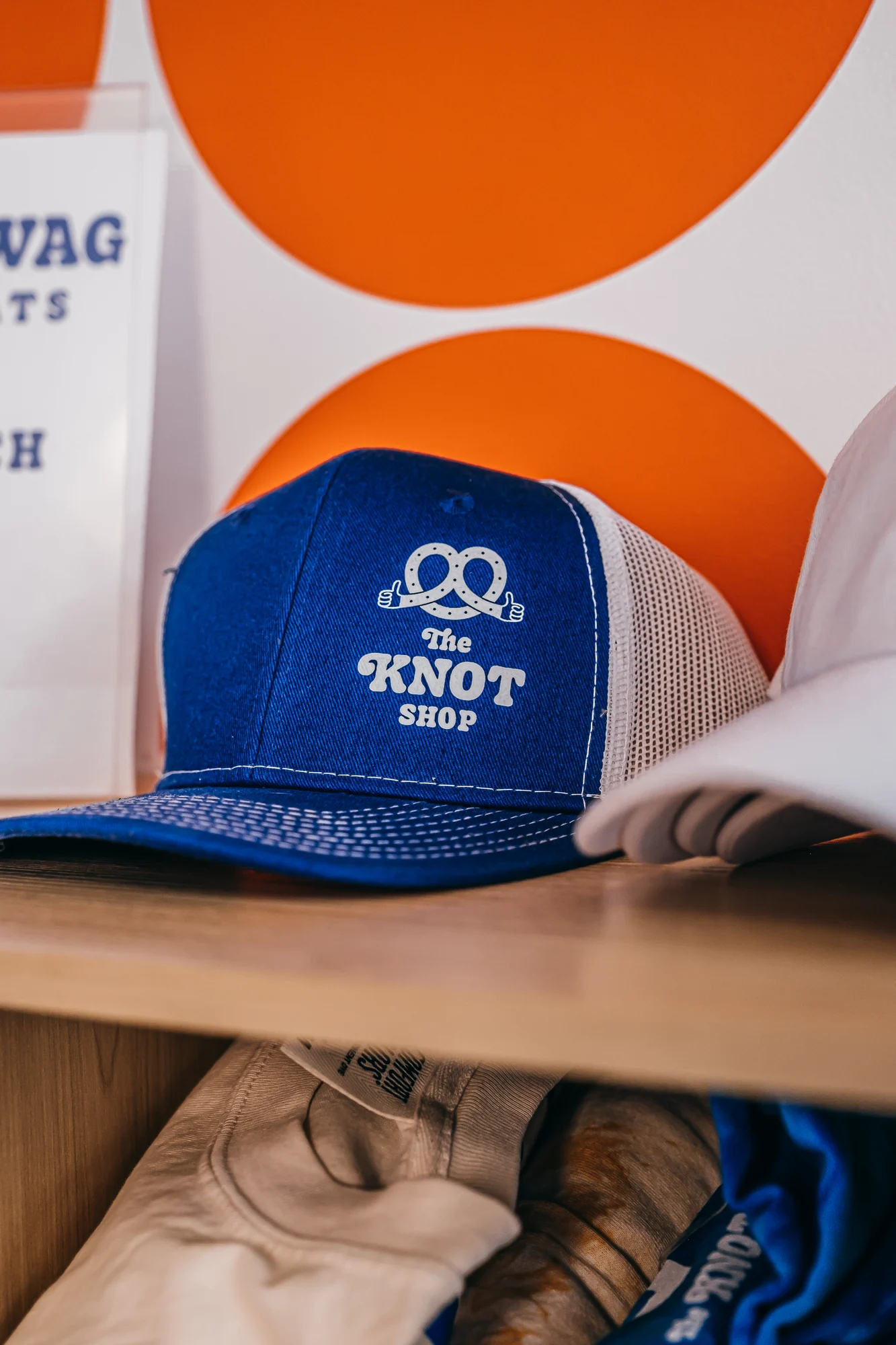 Blue mesh trucker cap with white knot graphic on wooden shelf and orange circles background.