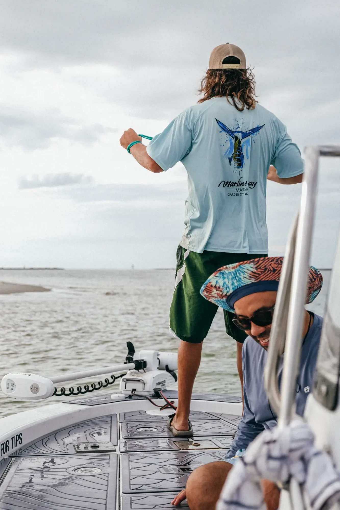 Two people on a boat by calm water under overcast sky one wearing marlin shirt.