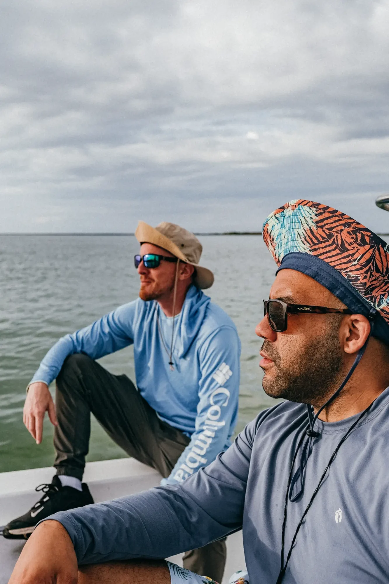 Two men wearing hats and sunglasses sit on a boat under a cloudy sky over calm water.