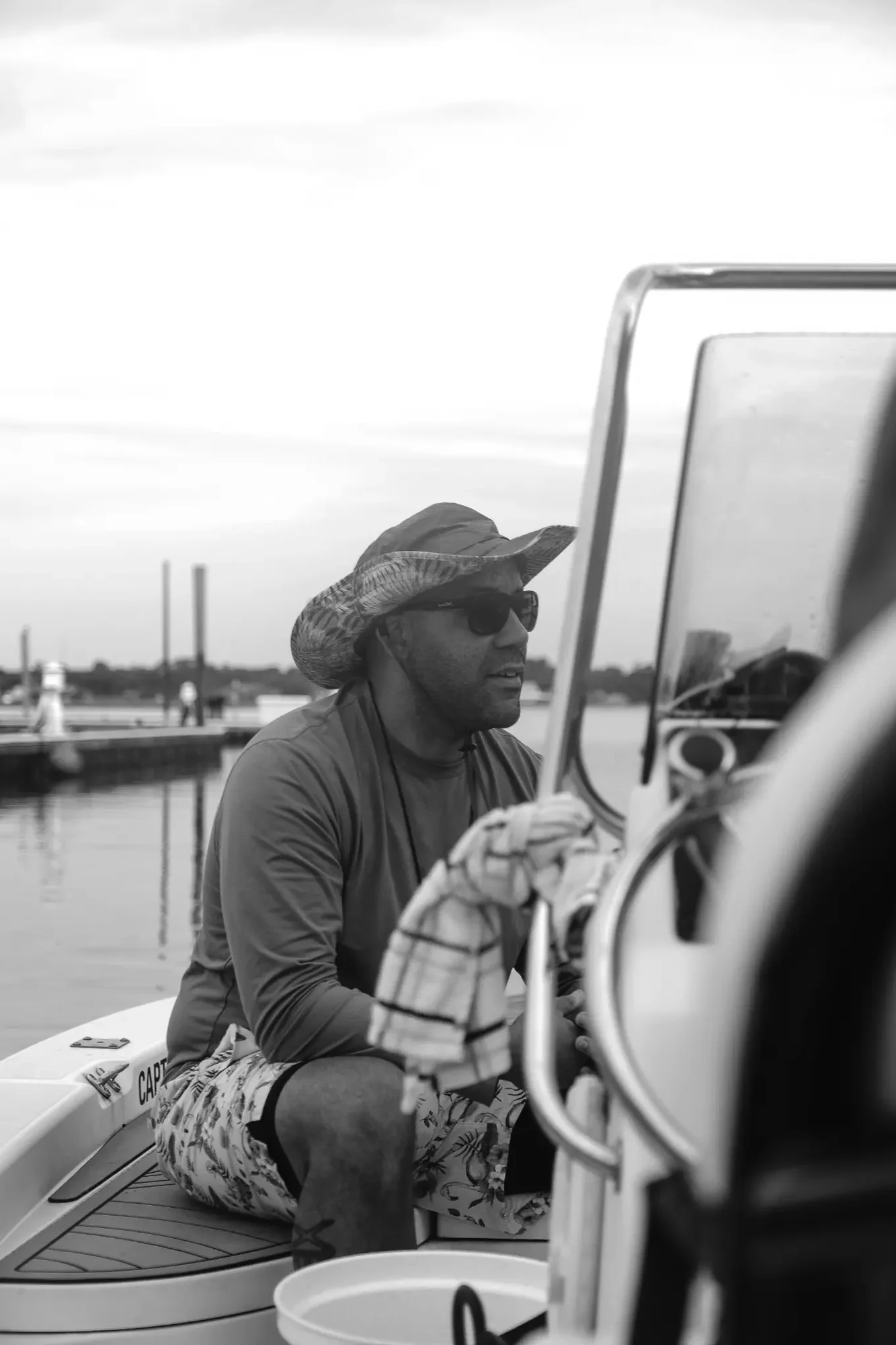 Black and white scene with person wearing wide brim hat and sunglasses seated in boat.