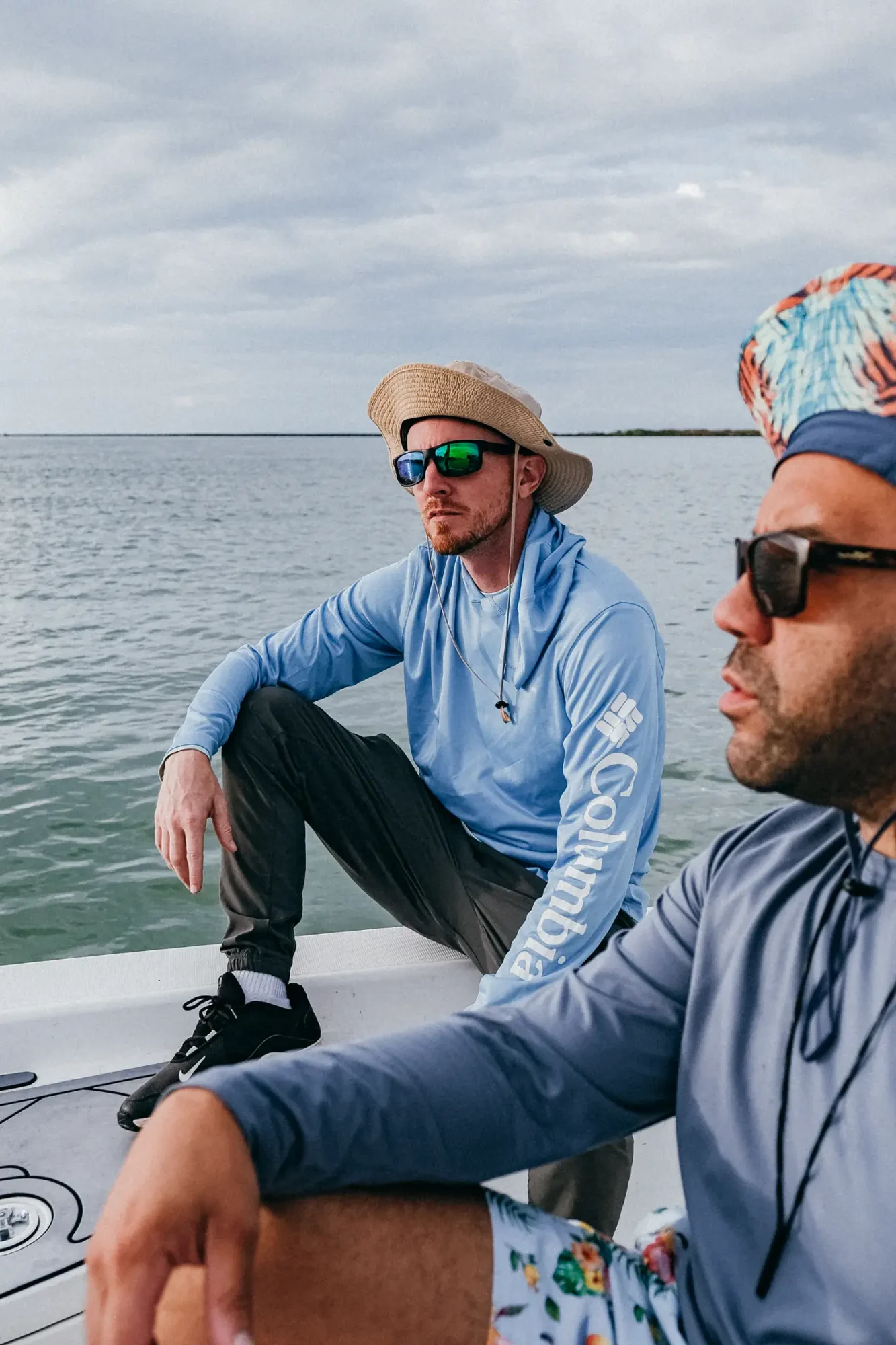 Two men wearing sunglasses with one in a straw hat seated on a boat over calm gray water.