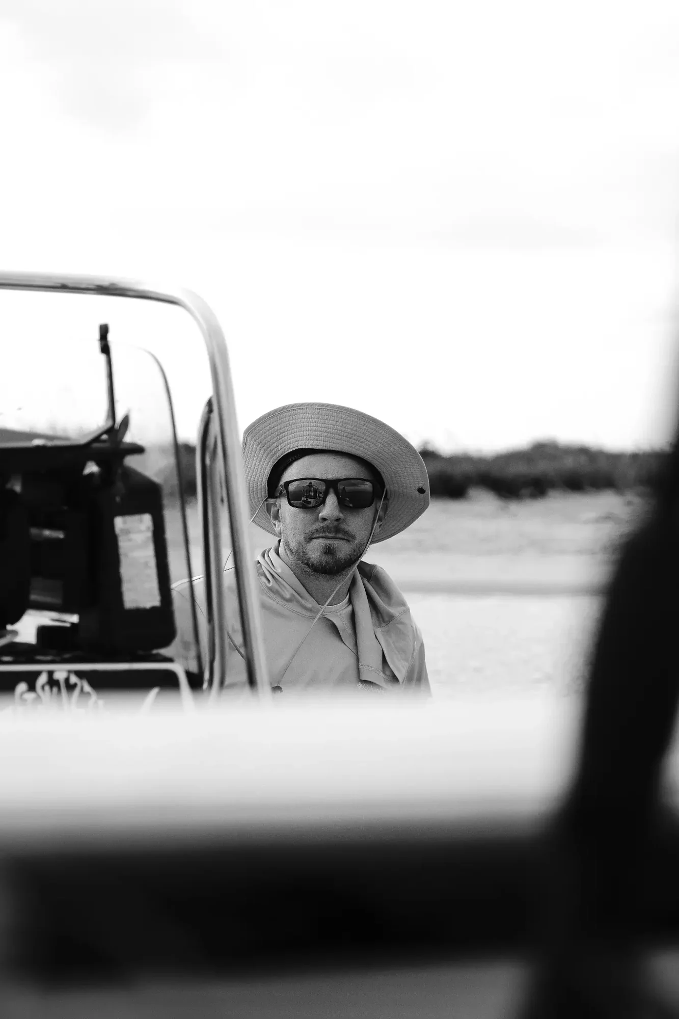 Man wearing wide brim hat and sunglasses beside boat with blurred shoreline background.