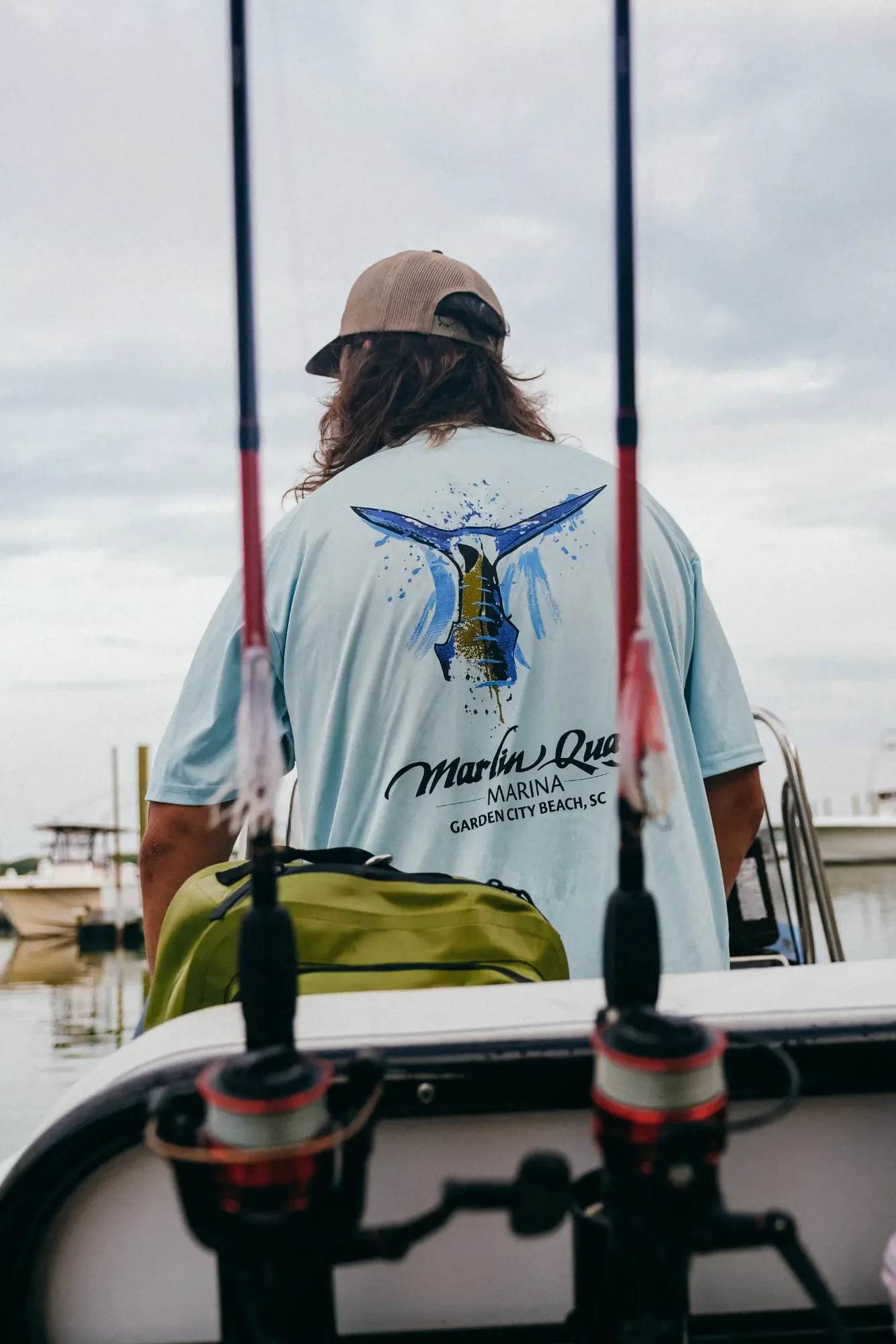 Person with long hair wearing beige cap and light blue shirt with fish graphic on boat.