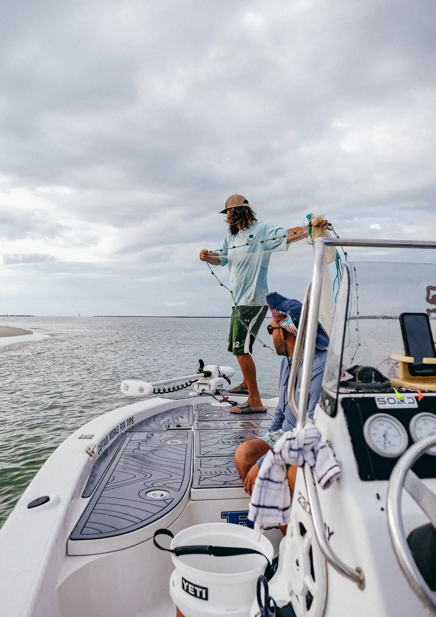 Person standing on boat bow casting net while another seated under cloudy gray sky.