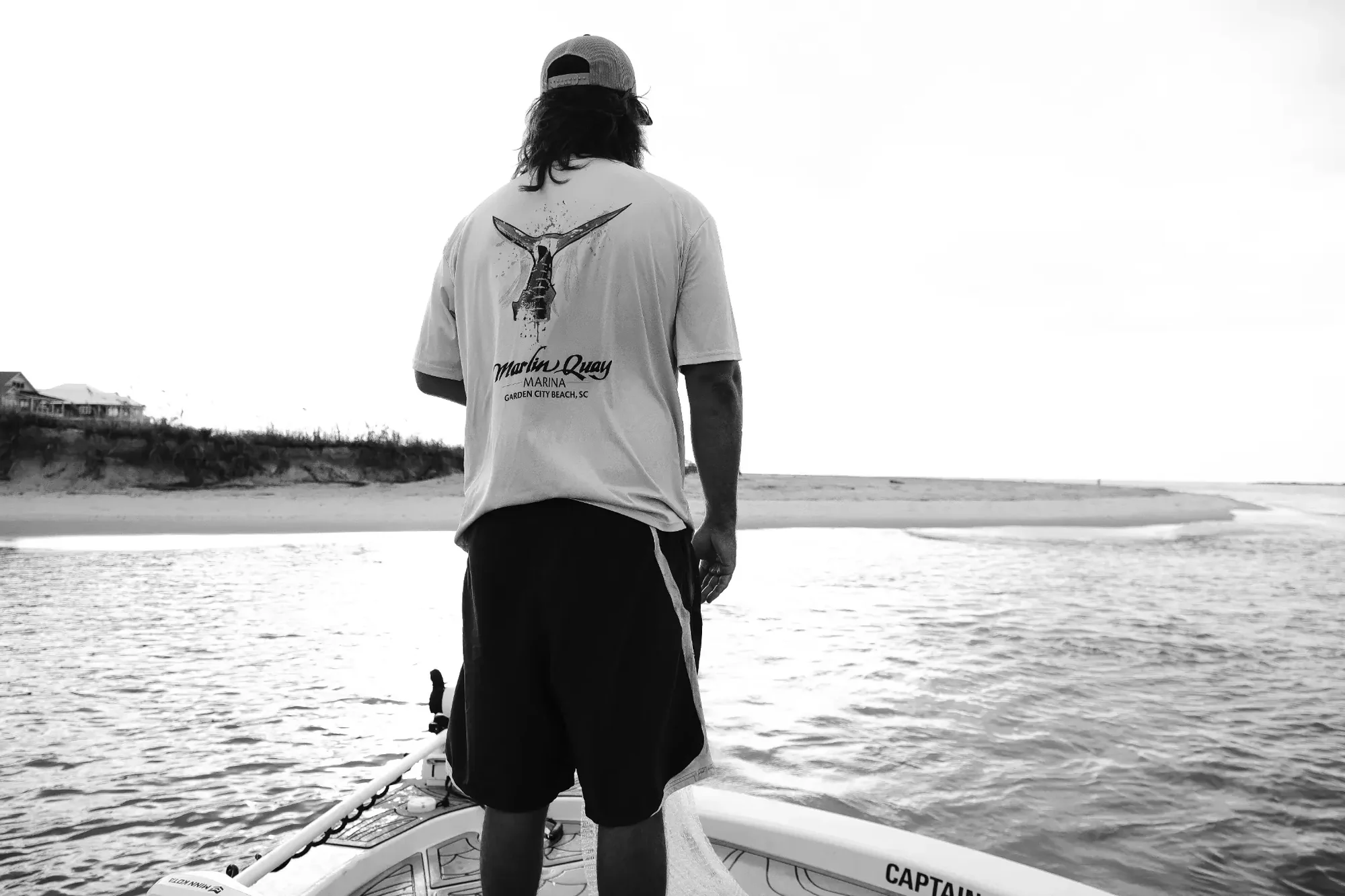 Backlit black and white scene of a person standing on a boat facing shoreline.