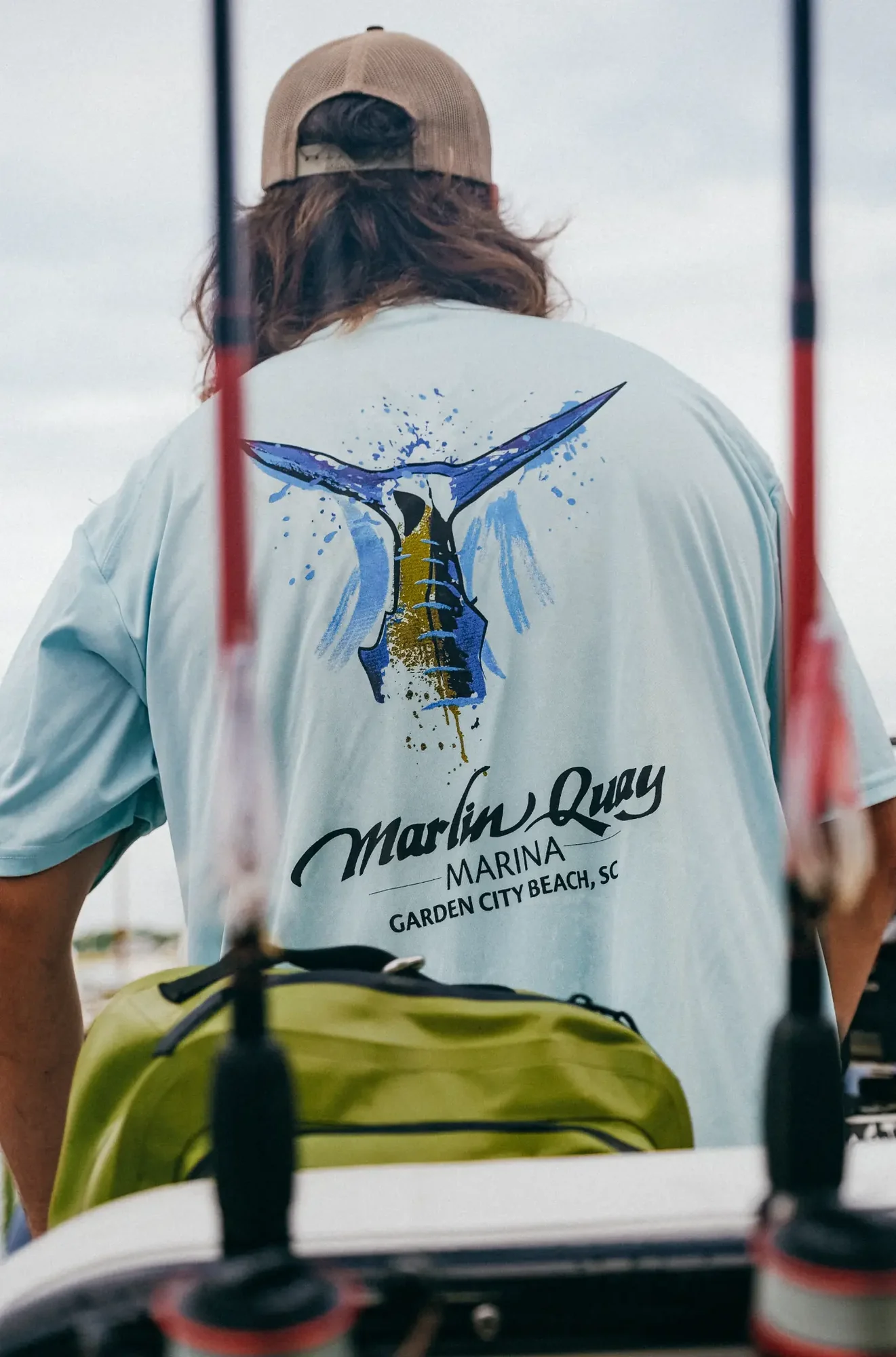 graphic marlin tail on light blue shirt worn by person facing away under overcast sky.