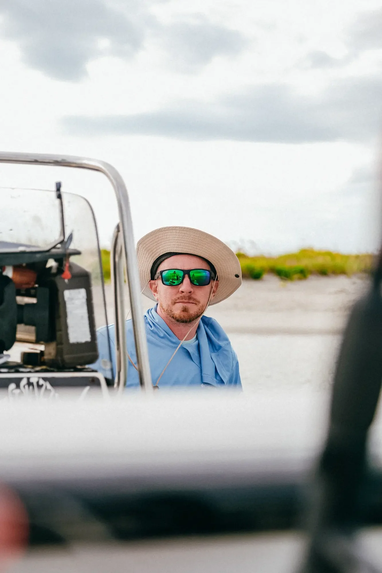 Person wearing wide brim hat and green mirrored sunglasses standing on boat under overcast sky.