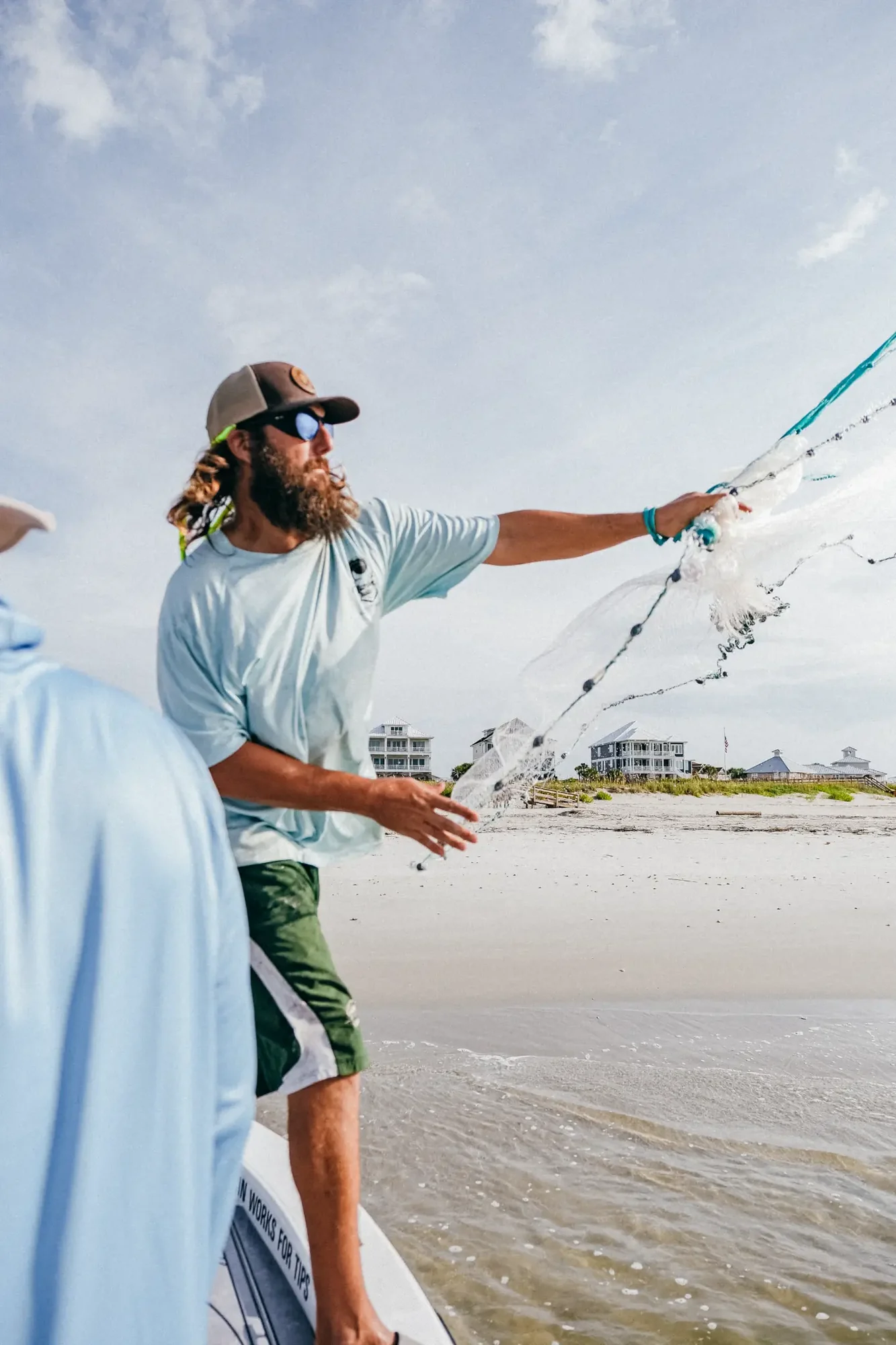 Bearded man in cap and sunglasses casting a fishing net from a boat on beach.