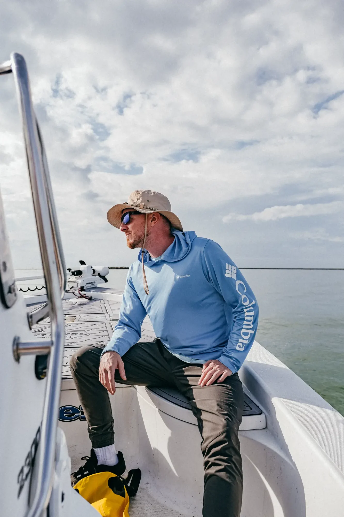 Man in tan bucket hat and blue hoodie sitting on boat under cloudy sky.