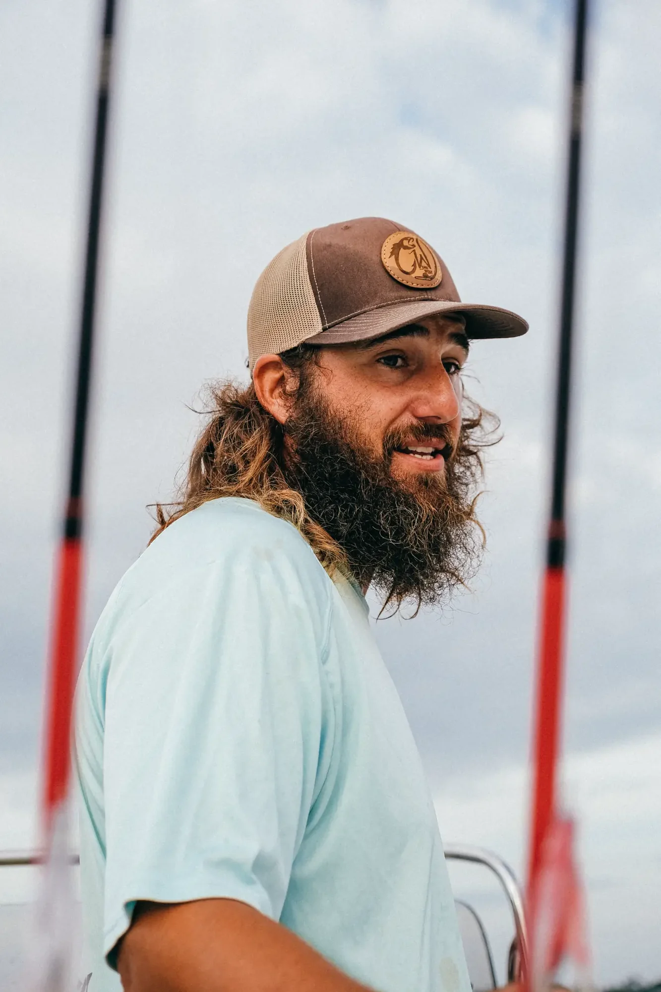 Bearded man in brown mesh cap and light blue shirt on boat deck under cloudy sky.