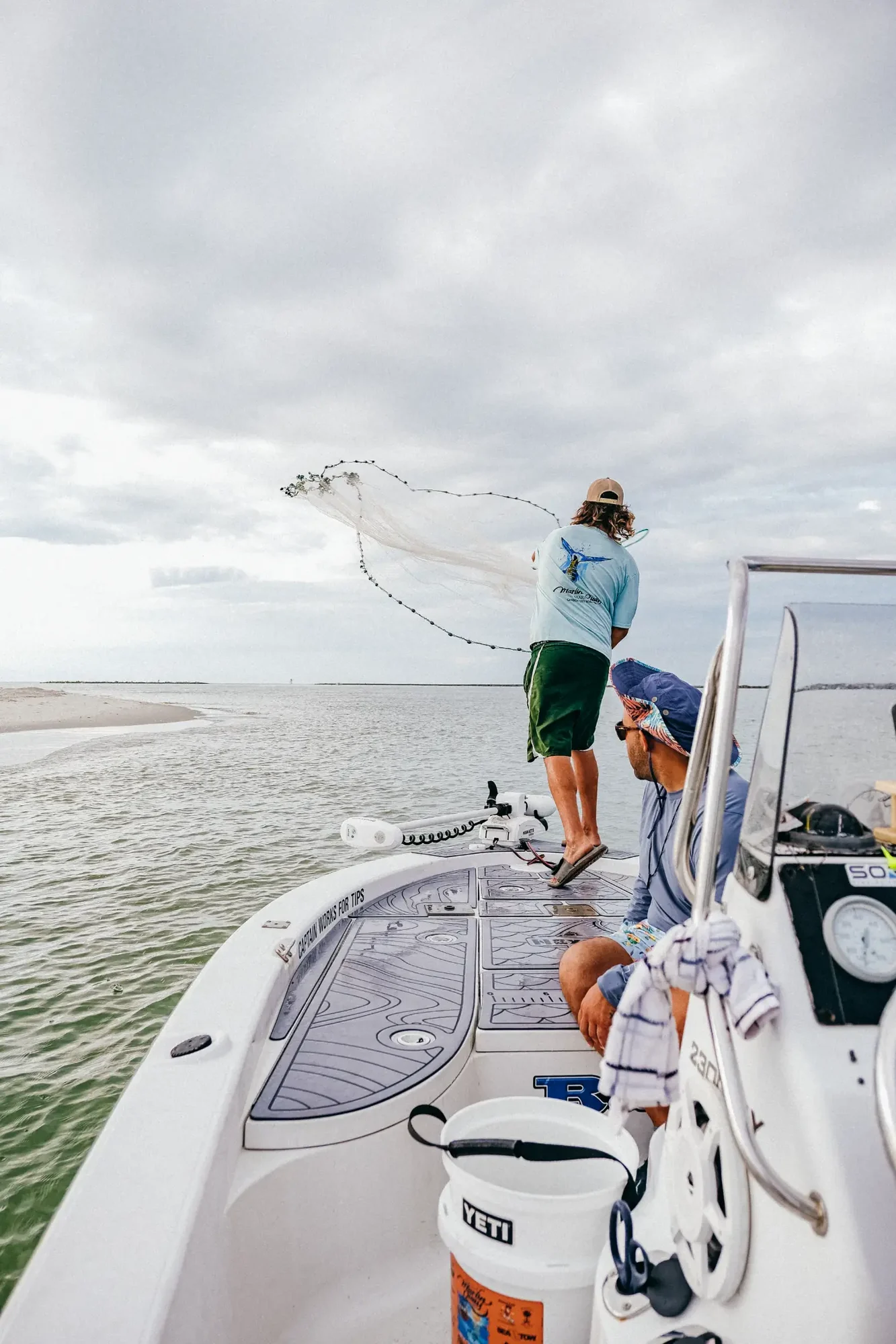 Person standing on boat casting circular net toward calm water under overcast sky.