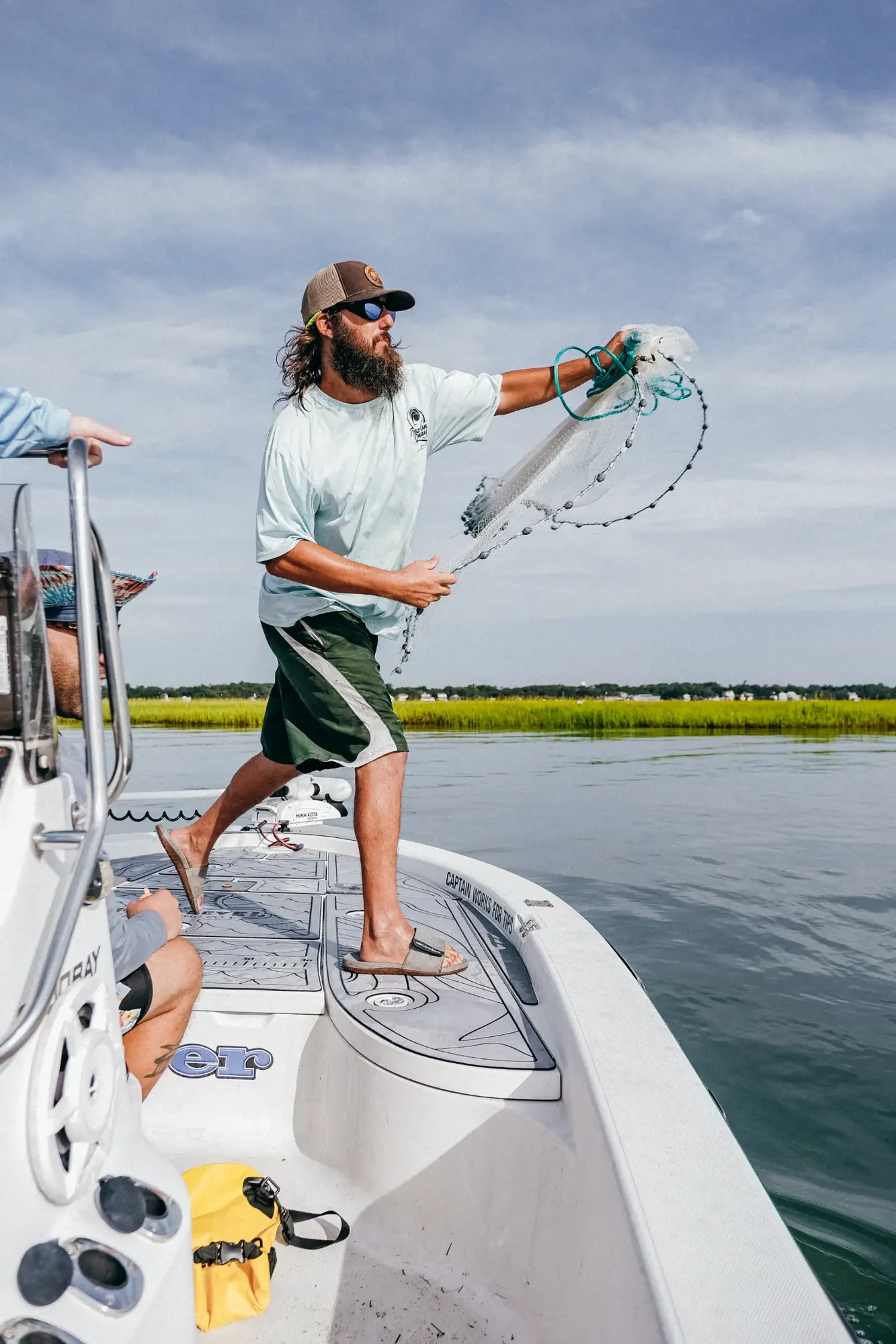 Bearded person in cap casting a net from boat over calm marsh waters.