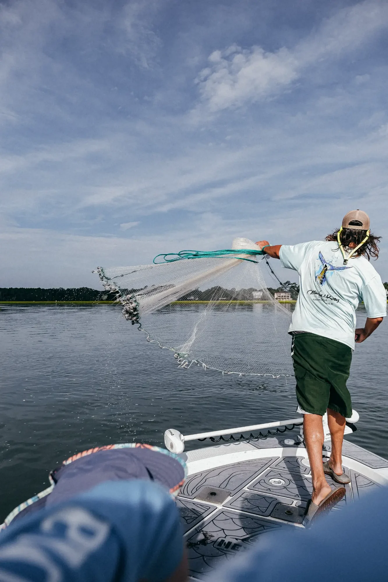 Person on boat casting a circular fishing net over calm water under pale blue sky.