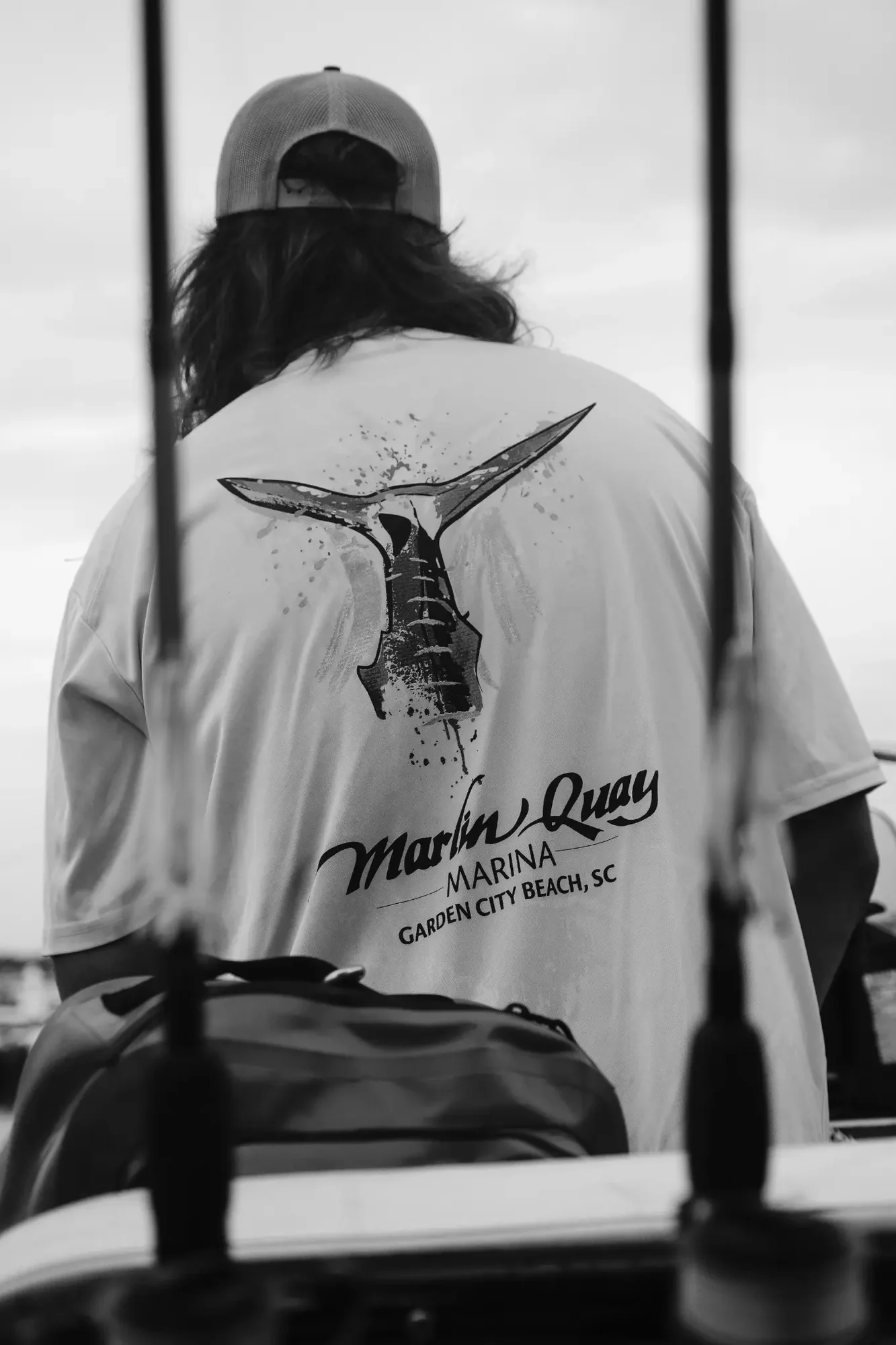 Black and white rear view of person wearing cap and shirt with marlin tail graphic.