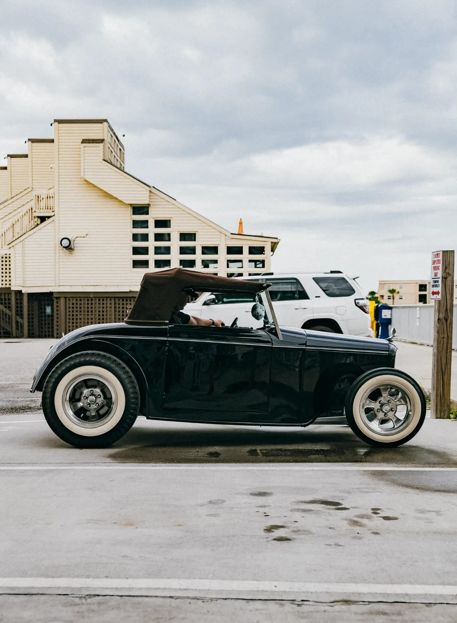 Black vintage convertible with whitewall tires parked before pale wooden building and cloudy sky.