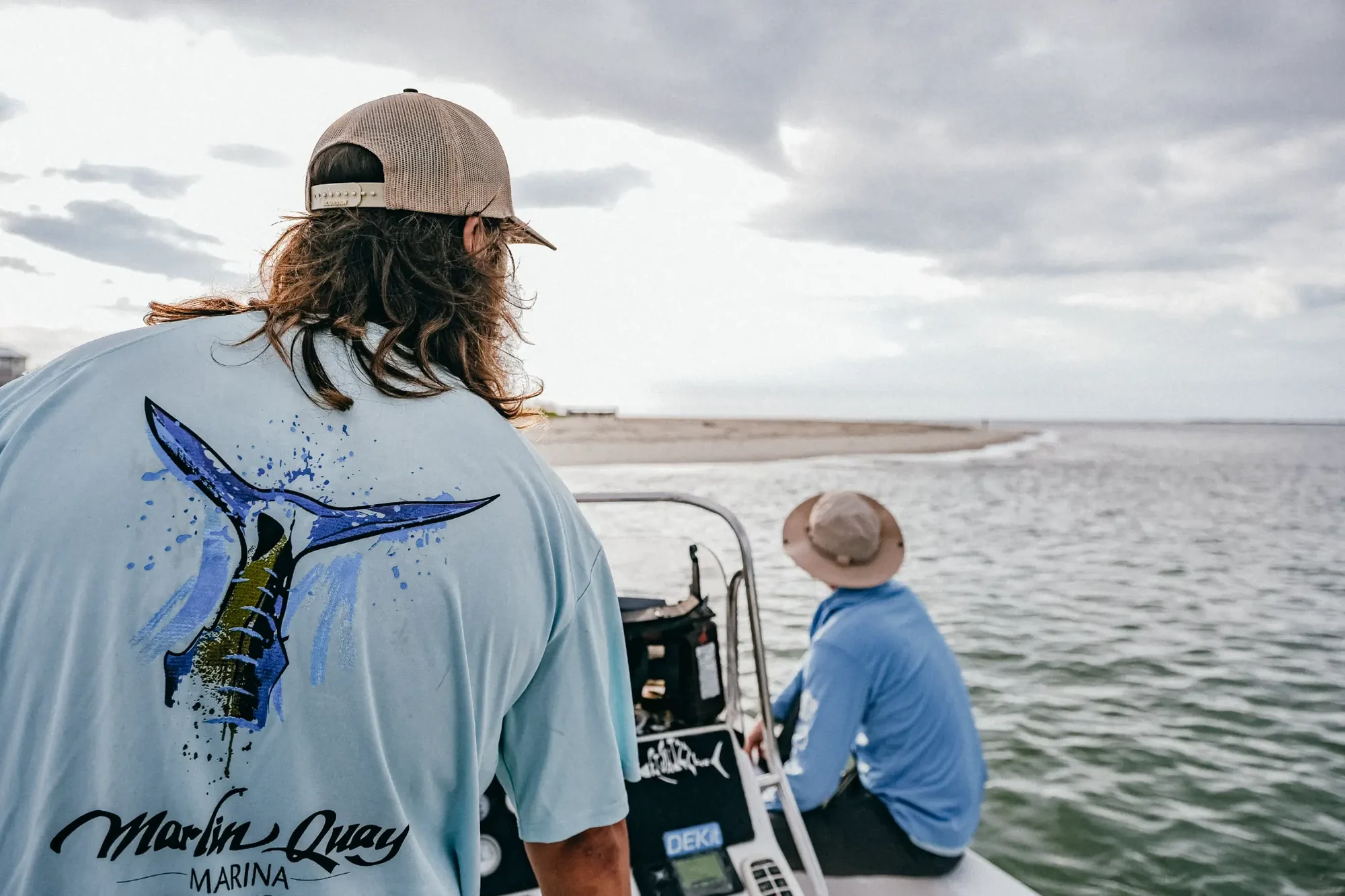 Person with long hair and cap in foreground wearing light blue fish graphic shirt on boat near cloudy water.