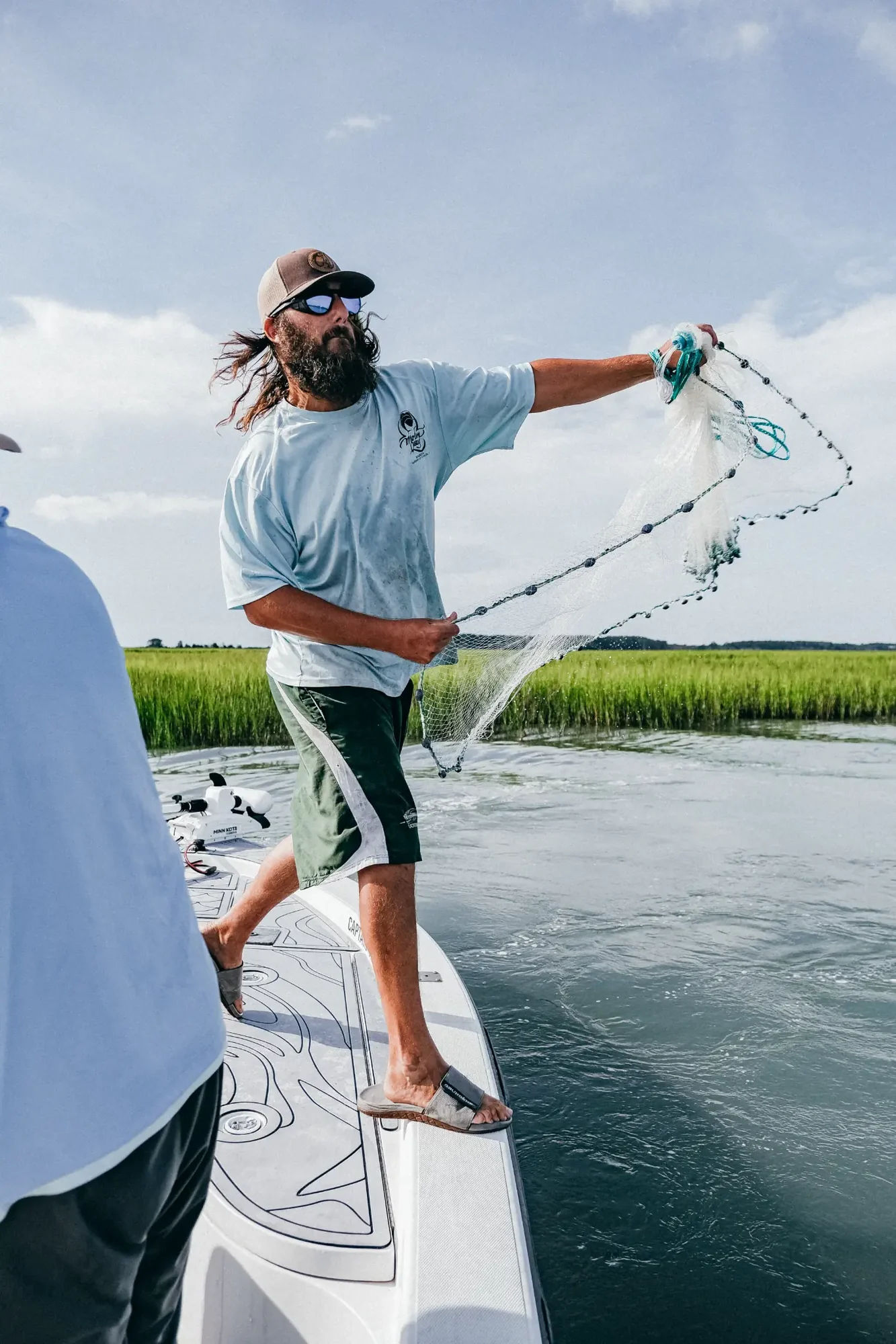 Bearded man on boat casting translucent net toward marsh under bright blue sky.