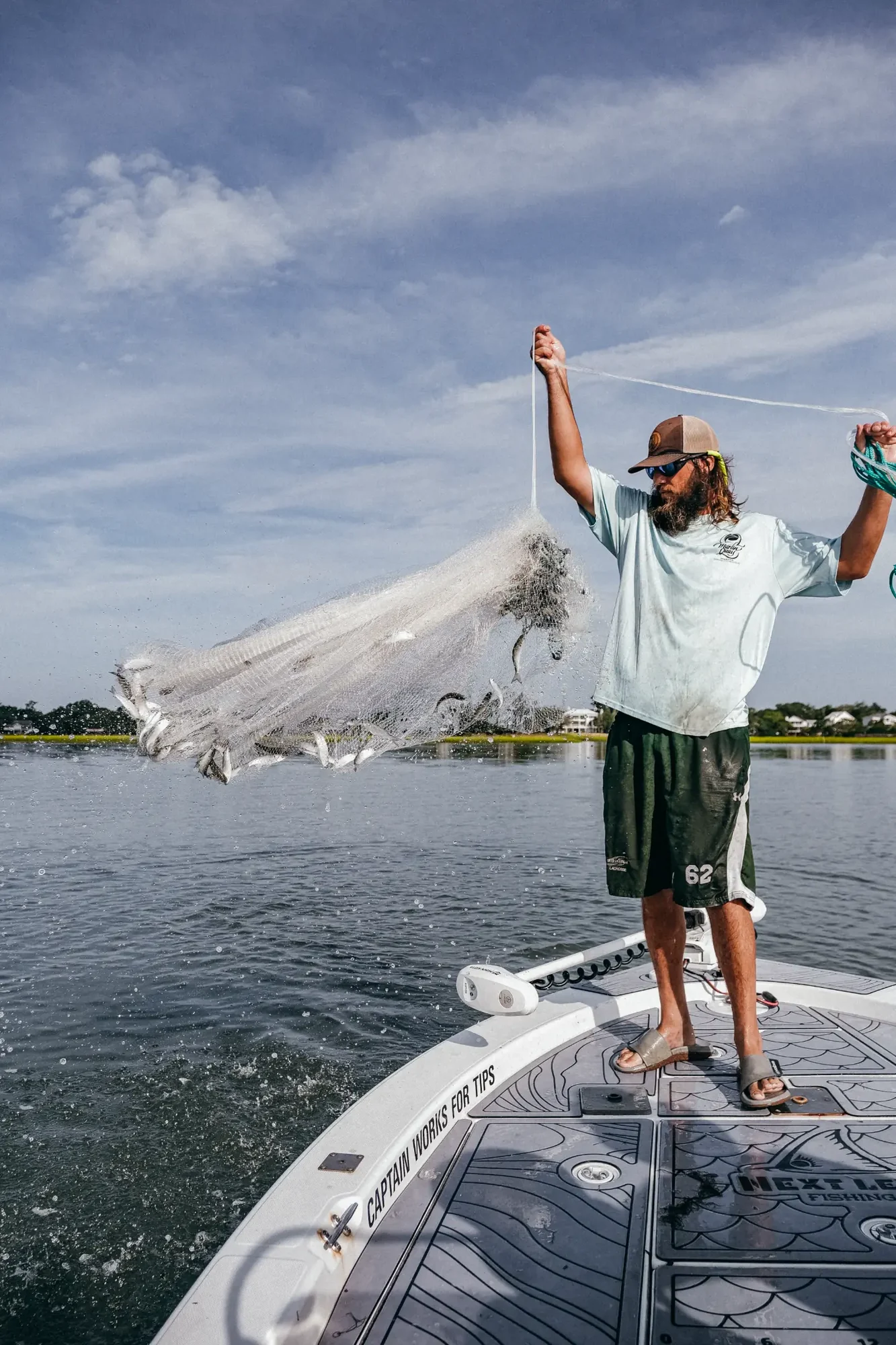Man on boat casting large fishing net across calm water under blue sky.