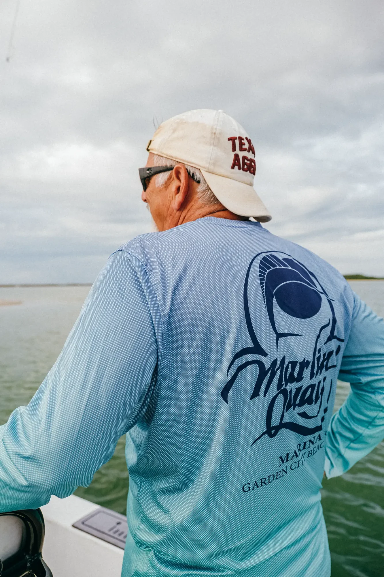 Man in light blue shirt and white cap worn backward leaning on boat railing over cloudy water.