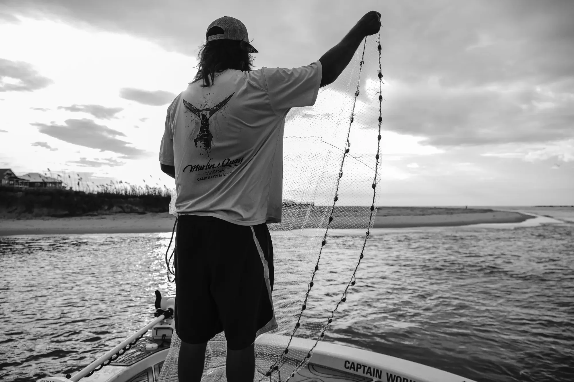 Black and white scene of person standing on boat holding net under backlit cloudy sky.