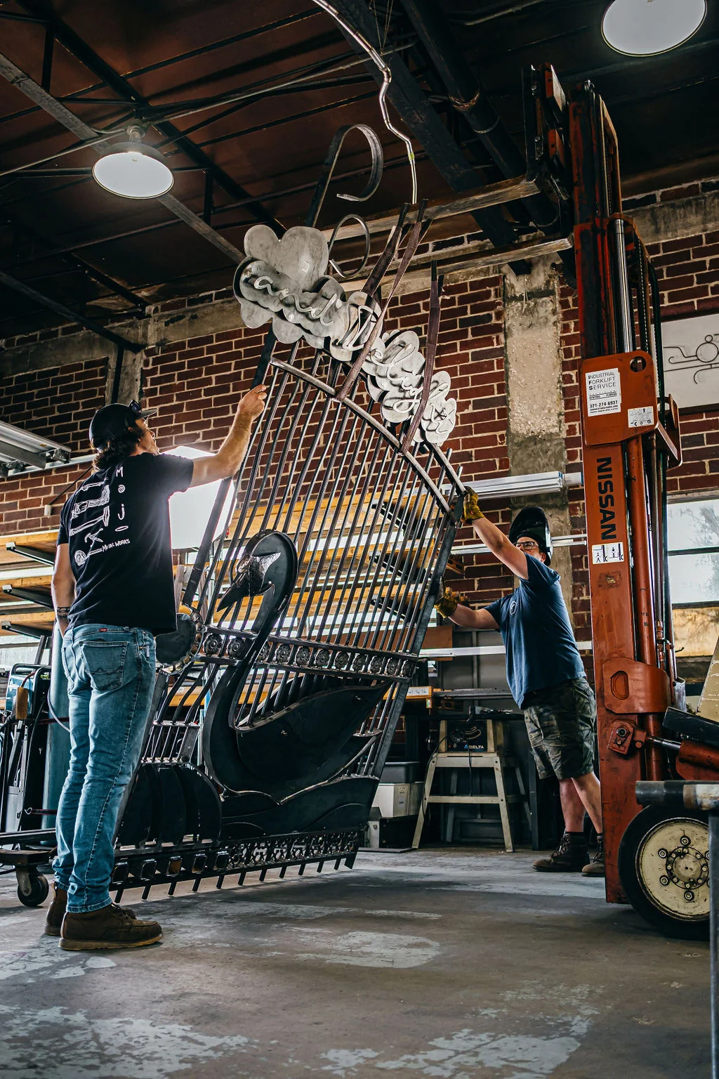 Two metalworkers maneuvering a tall welded animal sculpture inside the Mojo Metal Works studio.
