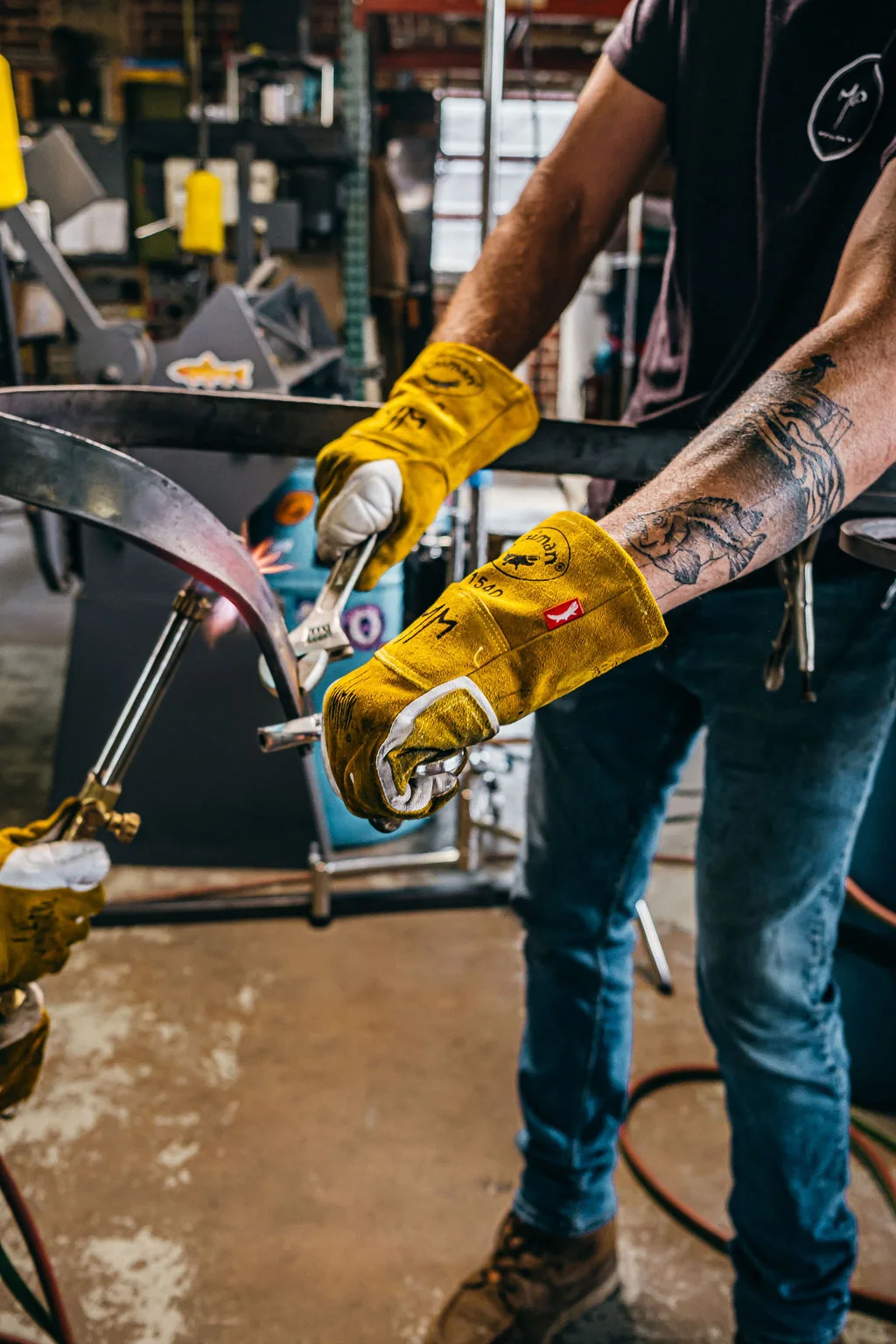 Tattooed forearms and yellow gloves guiding hot metal across a steel work surface.