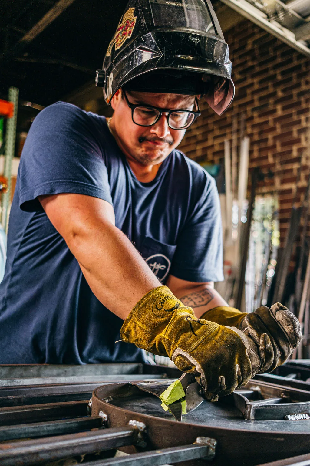 Metalworker in a welding mask shaping a curved steel form on a workshop bench.